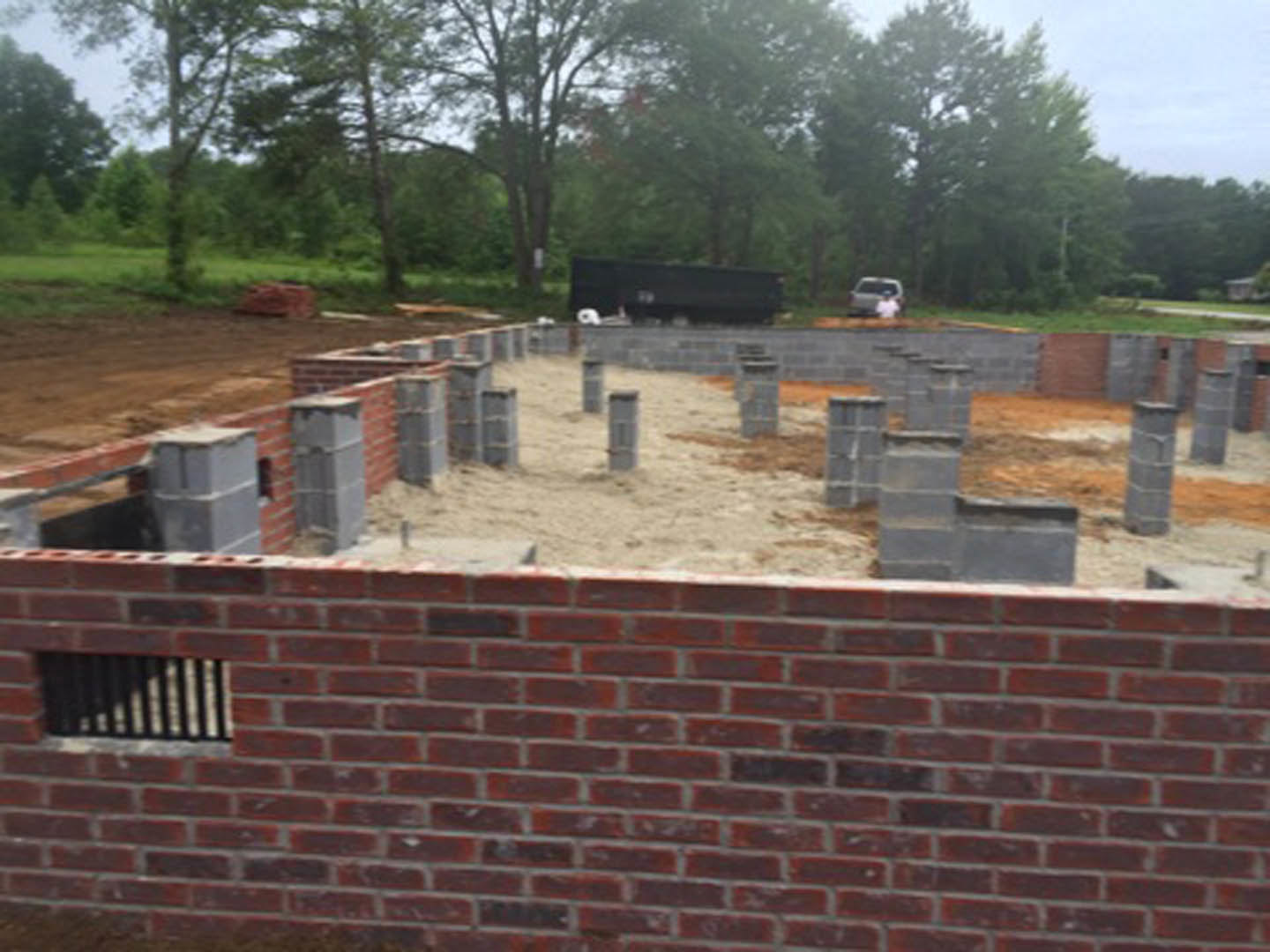 Red brick exterior wall with visible concrete foundation, dirt along the base, and tree branches in the background