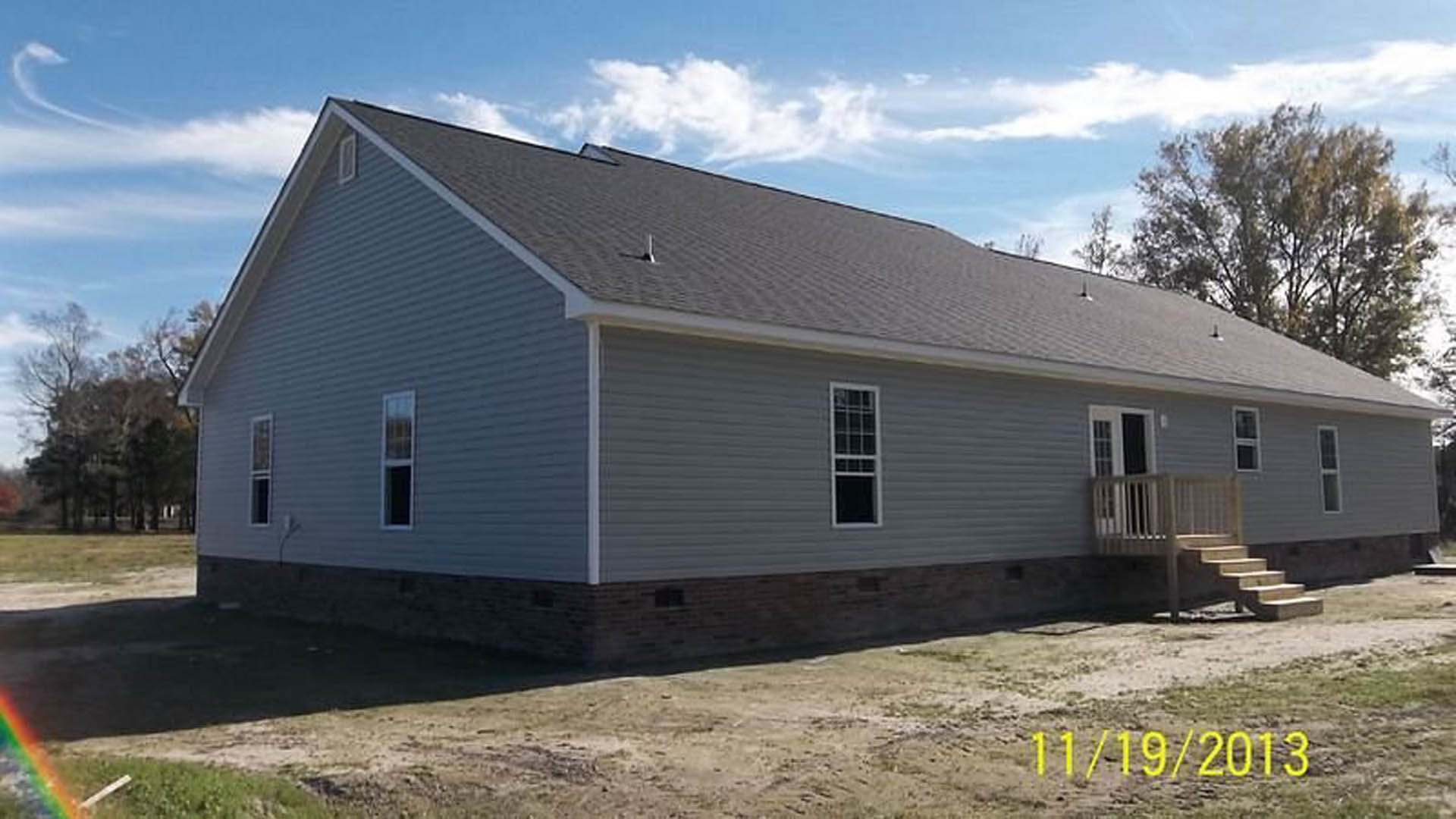 White-sided house with covered porch, multi-pane windows, and white support pole, set on snowy ground with Rockingham Meeting House visible in the background.