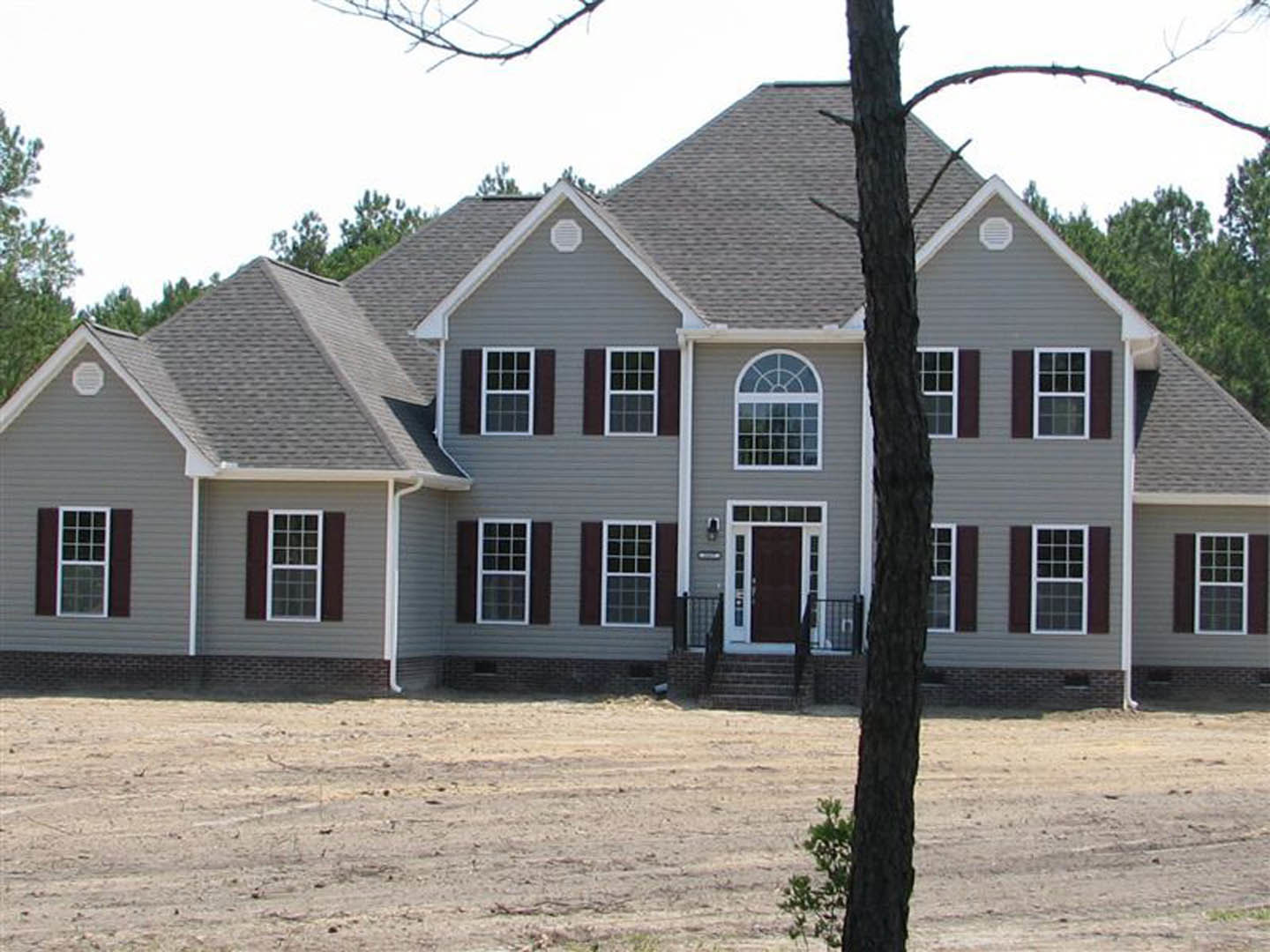 Grey house with black metal fence, tree in dirt yard, brick wall along property, close-up of window and door, horizontal siding and shingle roof visible