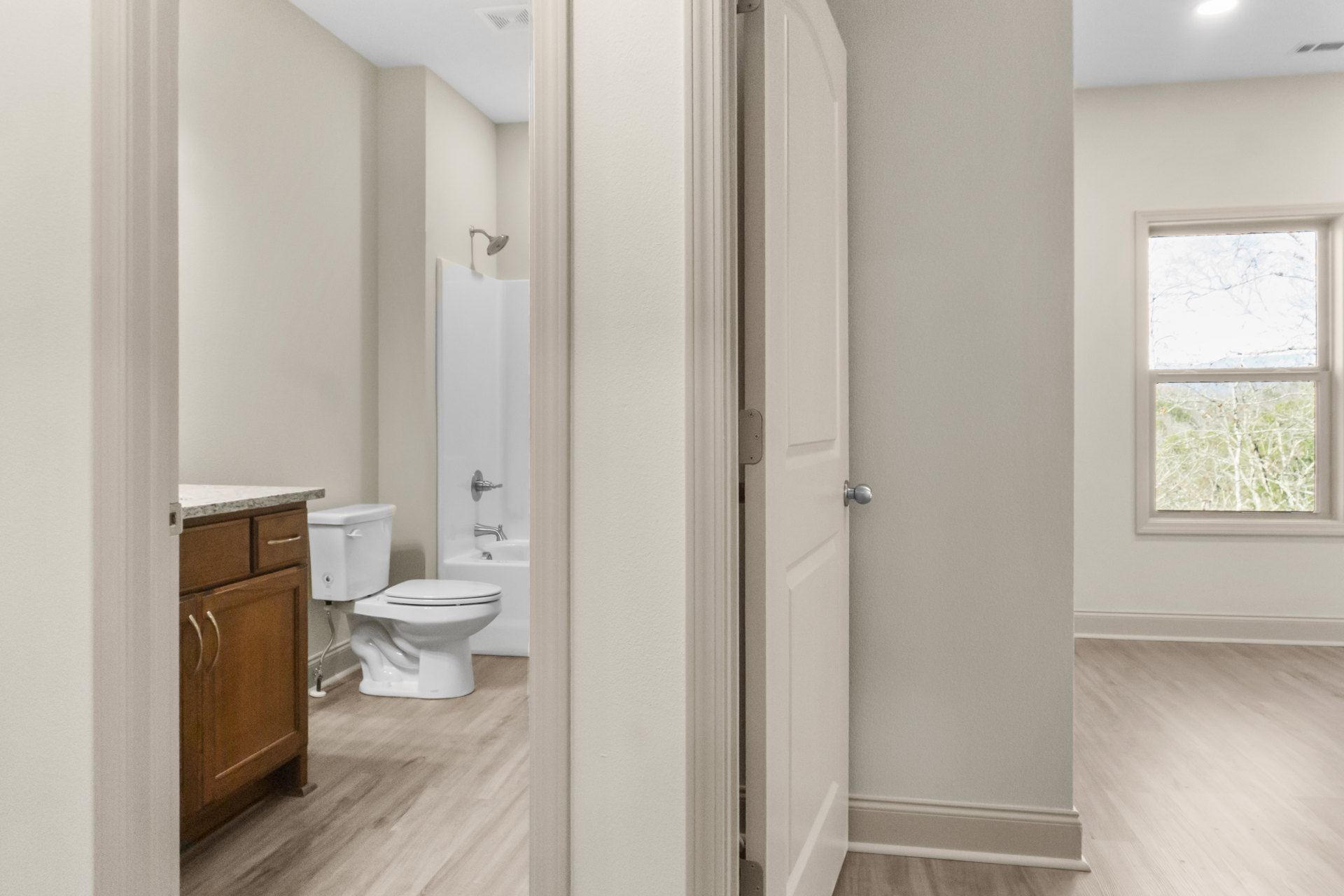 Bathroom with white toilet and rectangular sink, wooden vanity cabinet with metal handles, large window showing green trees outside, light tile flooring, plaster walls.