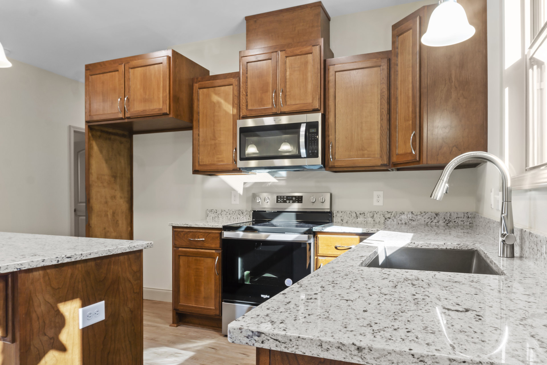 Granite countertops, wood cabinetry, stainless steel stove and oven, built-in microwave with illuminated glass, bell-shaped pendant light, sink, and white outlet on kitchen counter