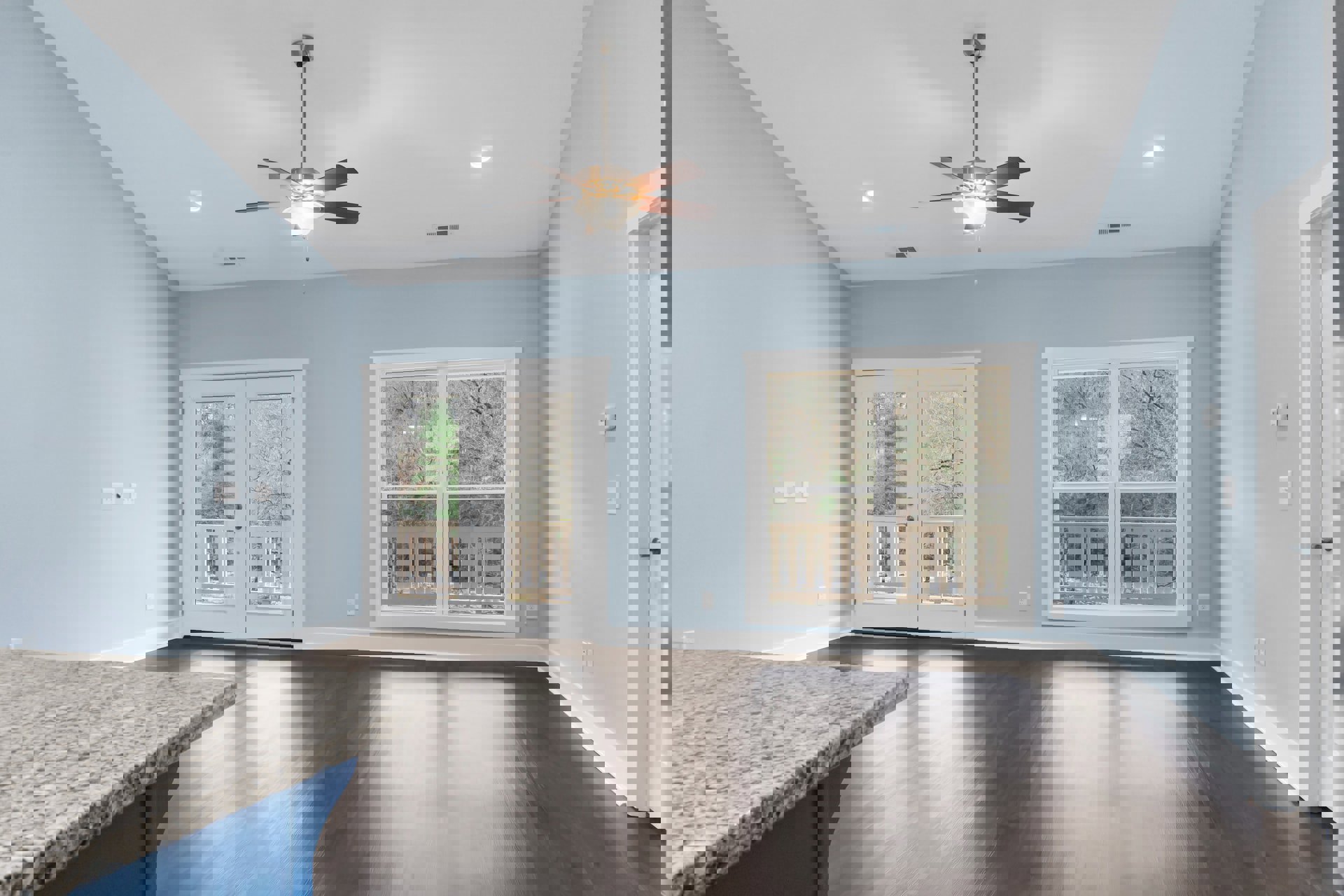 Open-concept room featuring a marble countertop, white double doors with glass panes, wooden flooring, white walls, ceiling fan with light, window with railing and view of trees.