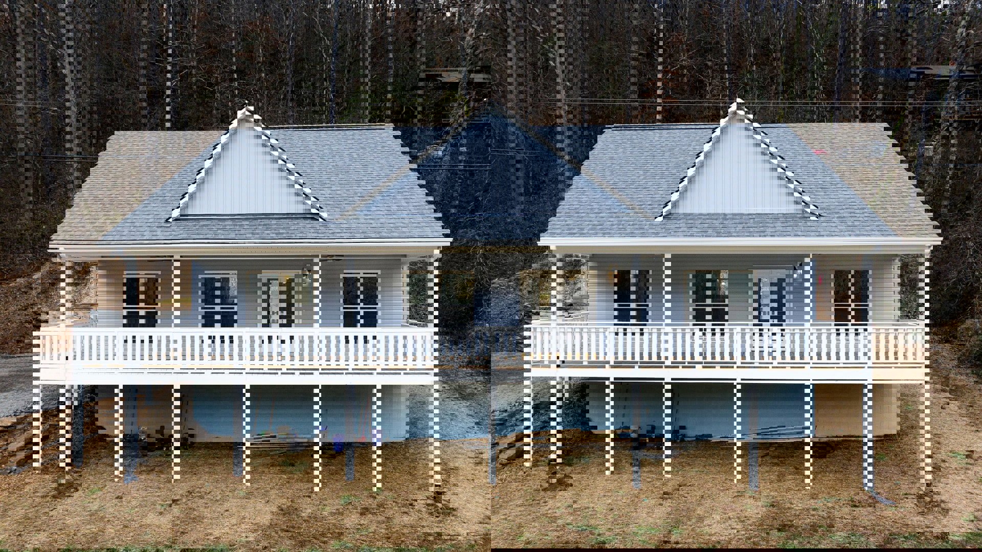 White cottage-style home with gray siding, covered front porch, elevated deck with white railing, white-framed windows, white entry door, and stacked lumber on the lawn.