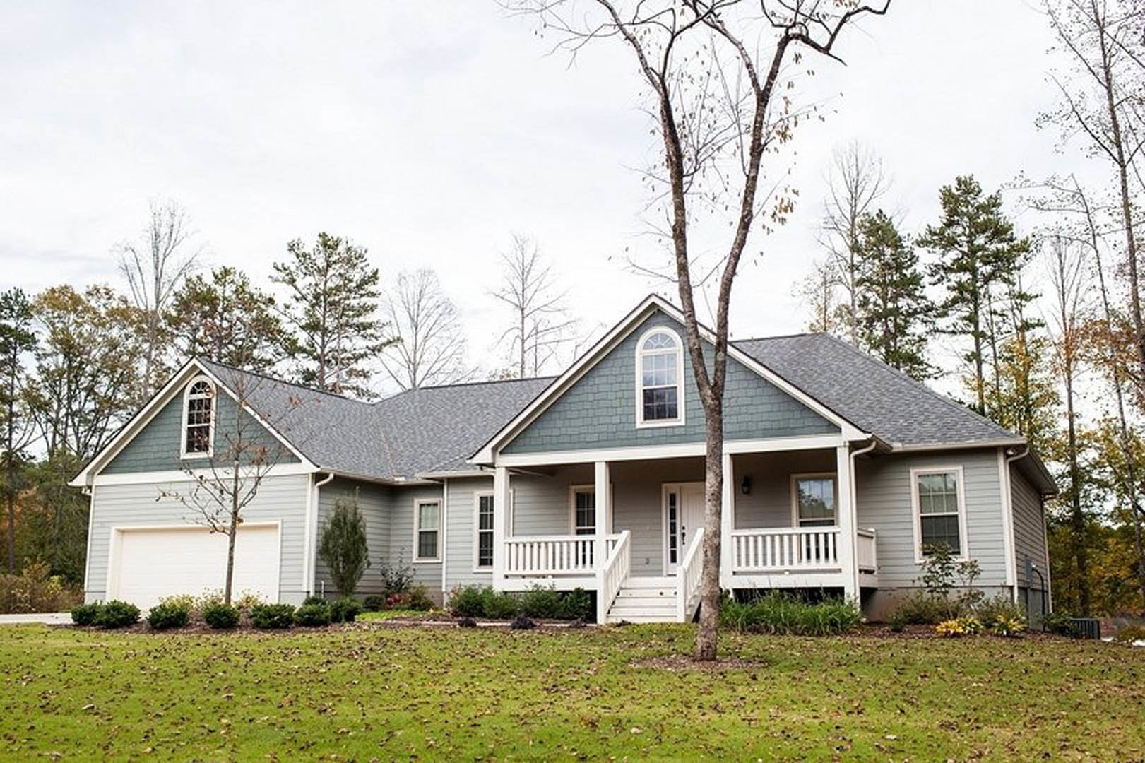 White cottage-style house with multi-pane windows, covered front porch, manicured green lawn, mature trees, and landscaped garden beds