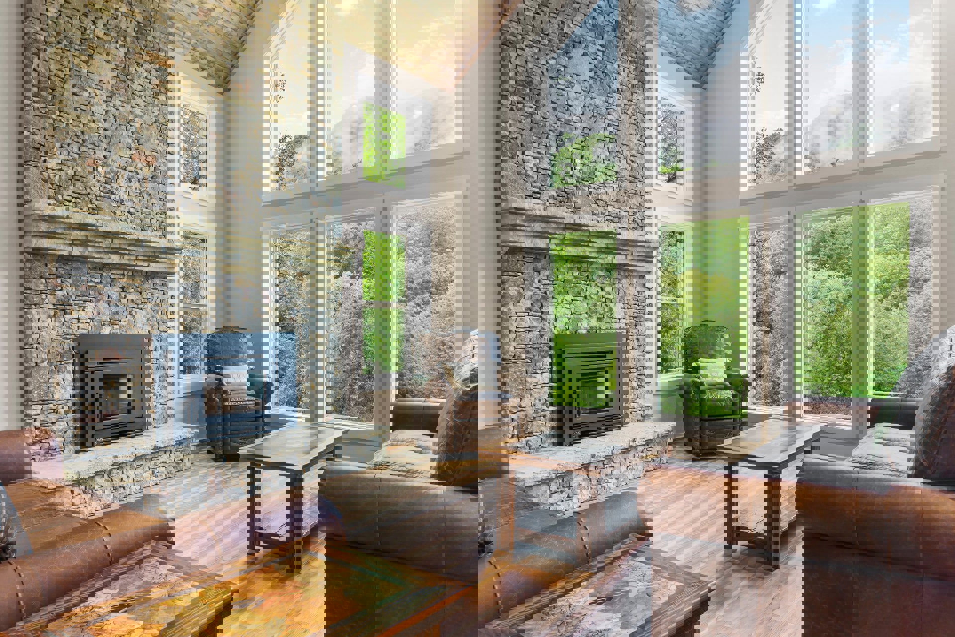 Living room with stone fireplace, brown leather chair with patterned pillow, gray sofa in front of large window, wood coffee table on light hardwood floor