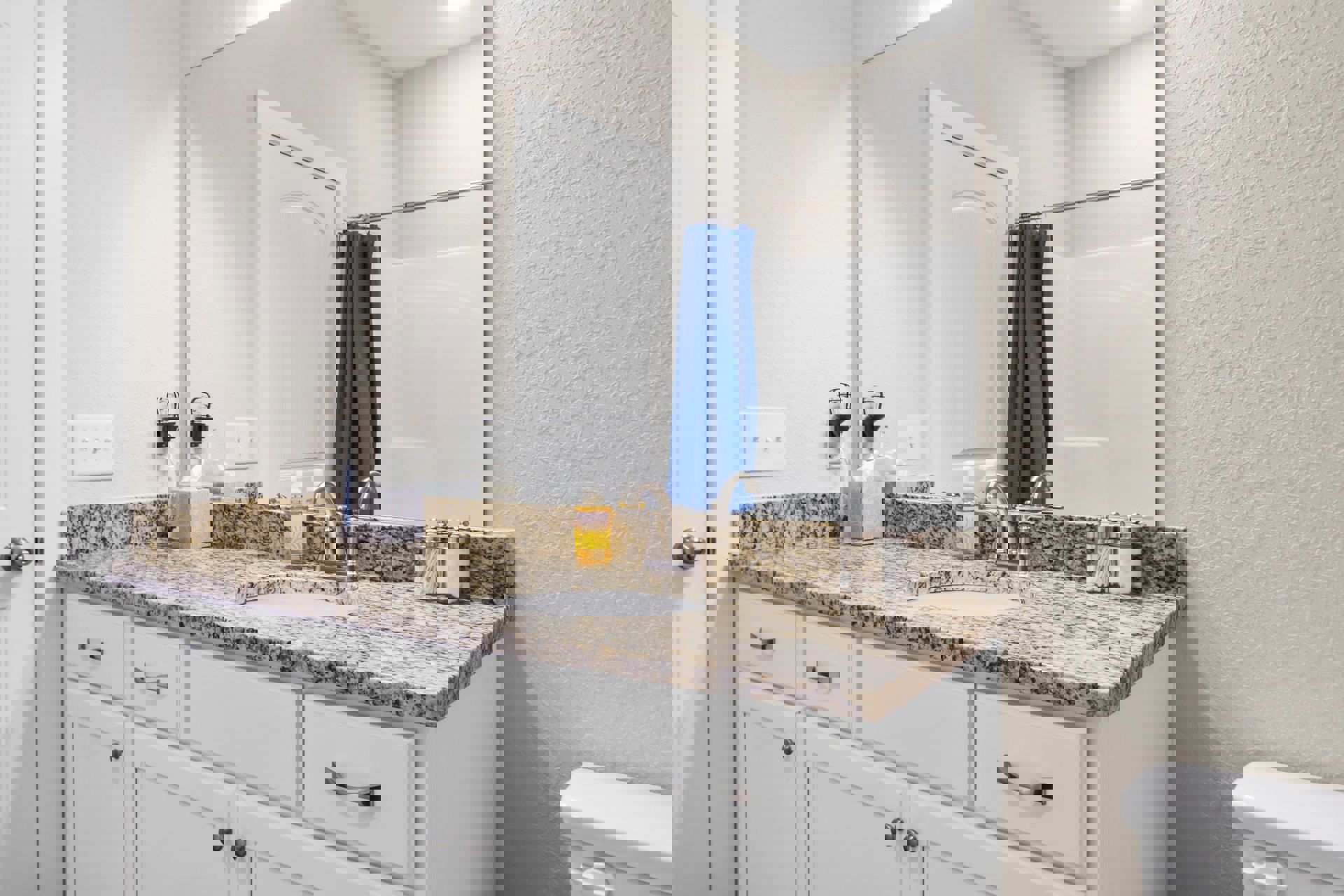 Marble bathroom countertop with a rectangular mirror, white toilet tank and handle, blue towel hanging from white door, blue curtain against white wall, three-switch white light