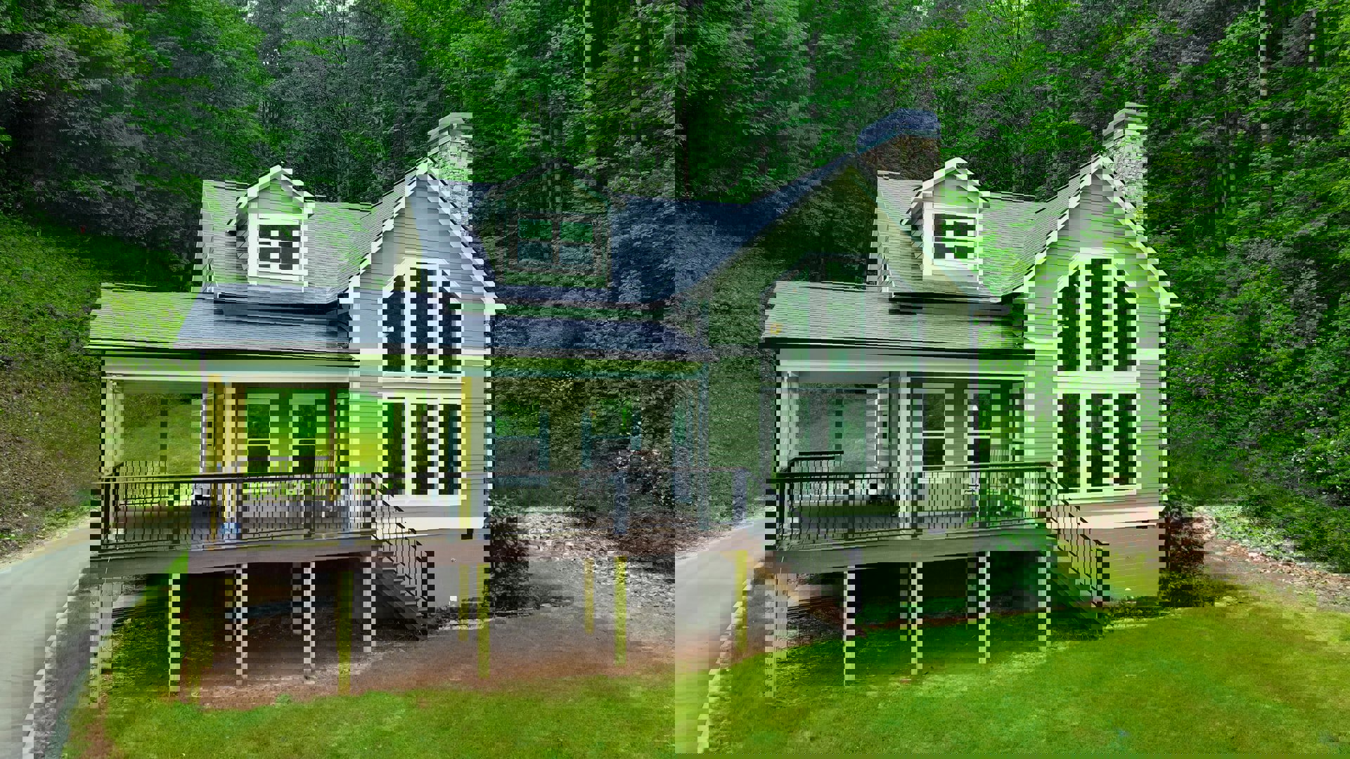 Two-story home with wood deck and white-railed porch, large windows, green bushes, and mature trees in landscaped yard