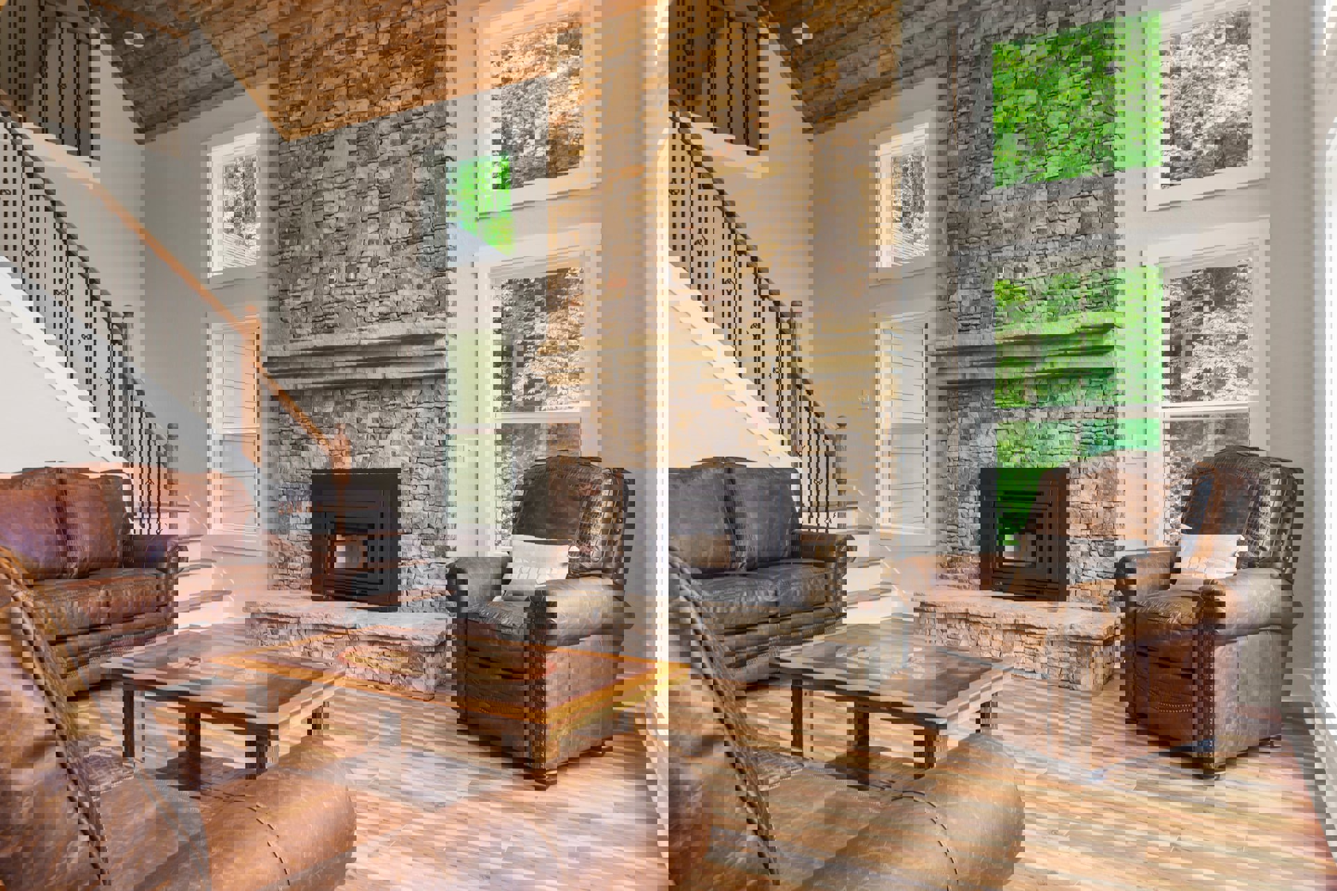 Brown leather couch and chair with accent pillows arranged around a black glass-door fireplace, wood coffee table centered on hardwood floors, large window framing leafy trees