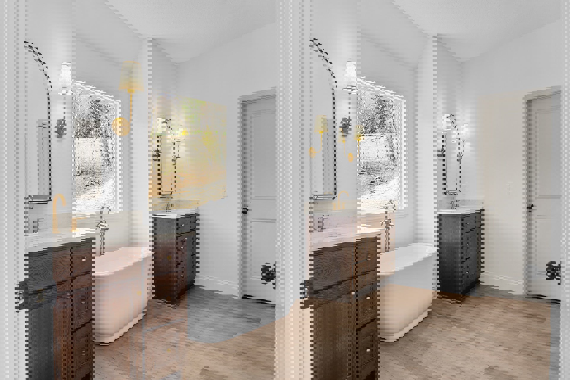 White freestanding bathtub on warm wood flooring beside a modern sink with chrome faucet, white lamp shade, and partial view of a house through window