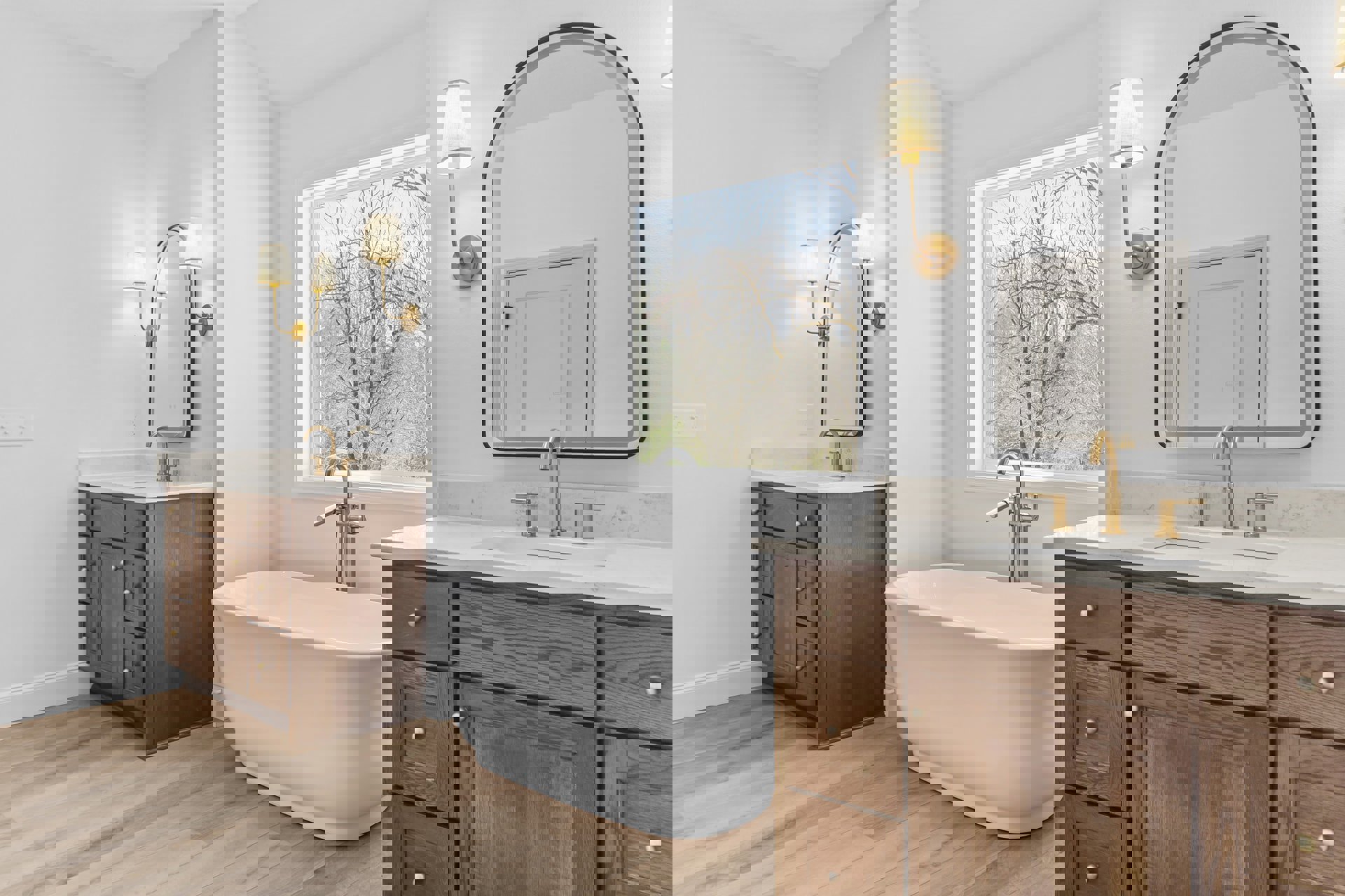 White freestanding bathtub beside a window with trees visible outside, white vanity with sink and gold faucet, wall mirror reflecting a white door with gold handle, lamp with white