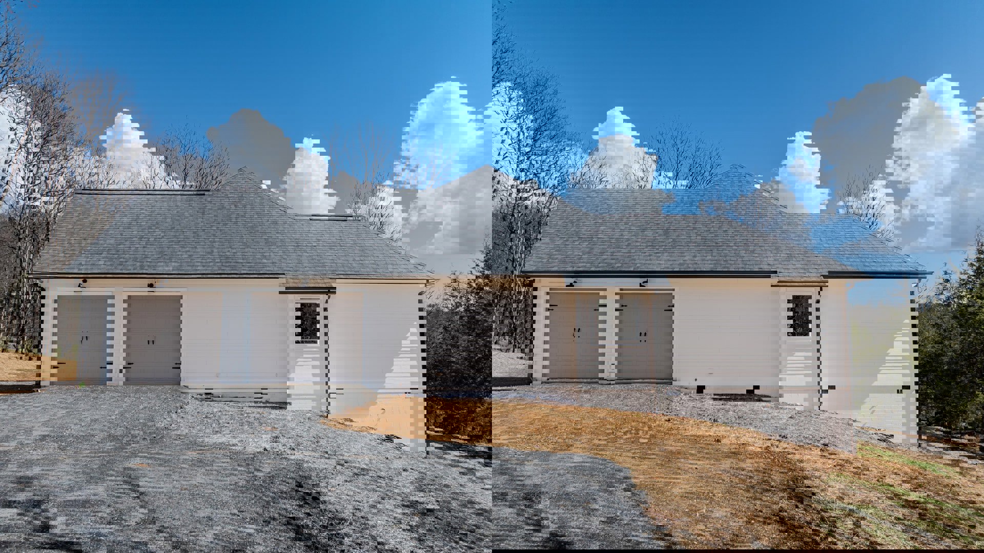 White house with gray garage door, black handles, white-framed window, dirt driveway, tree, and blue sky