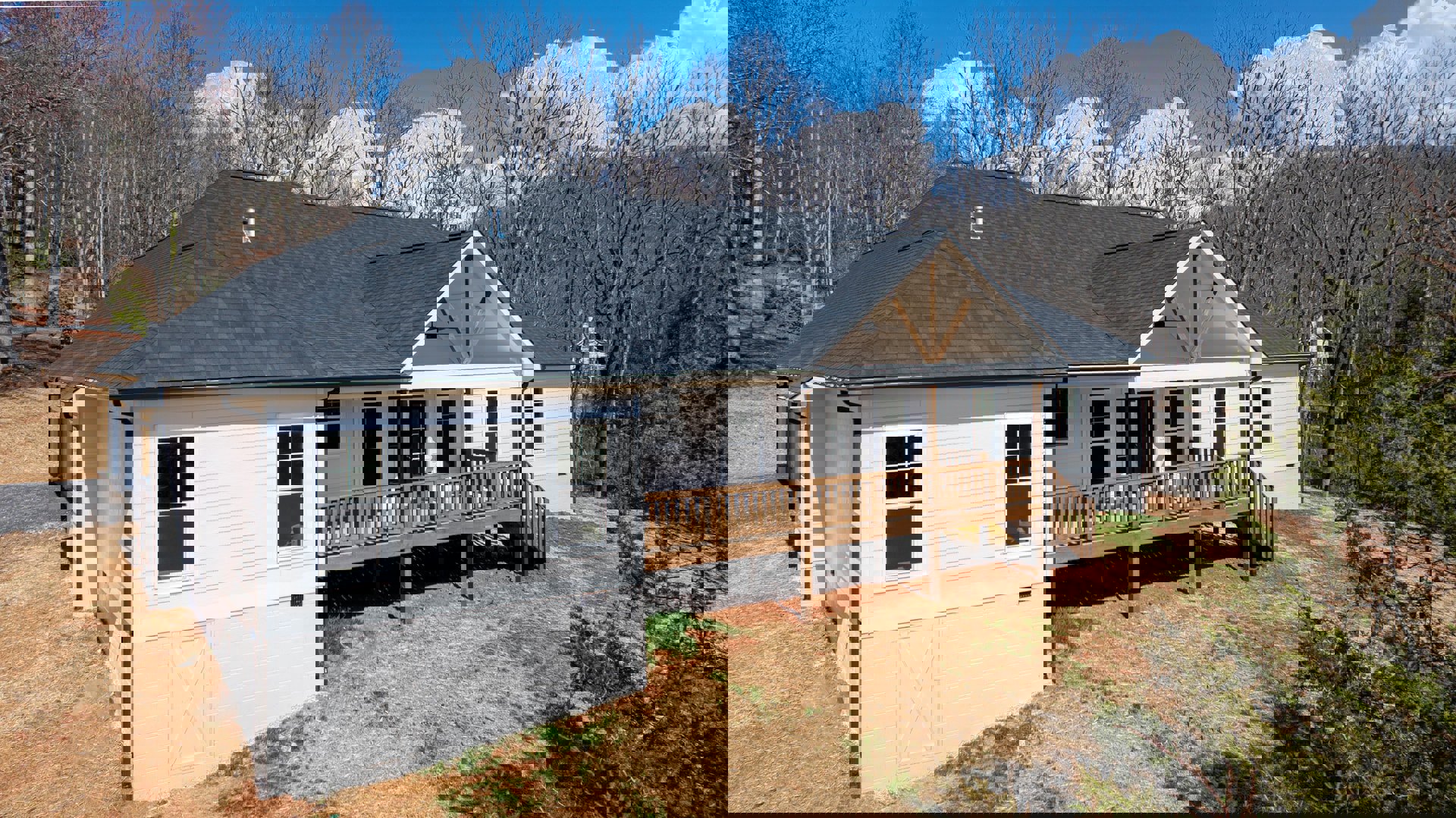 Wooden deck with railing attached to a cottage-style home, white-framed windows, covered porch, vented roof, surrounded by trees and sky