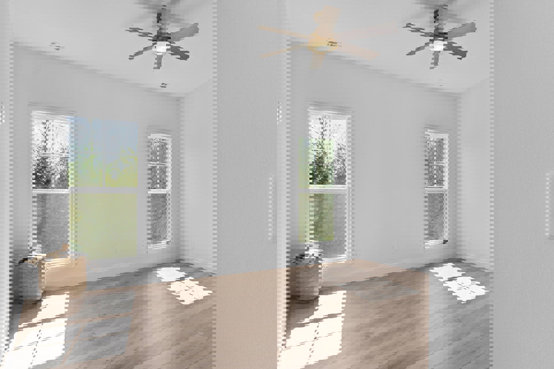 Wood flooring and white walls in a bright room with a ceiling fan, large windows framed in white, and a lidded basket; leafy trees visible outside.