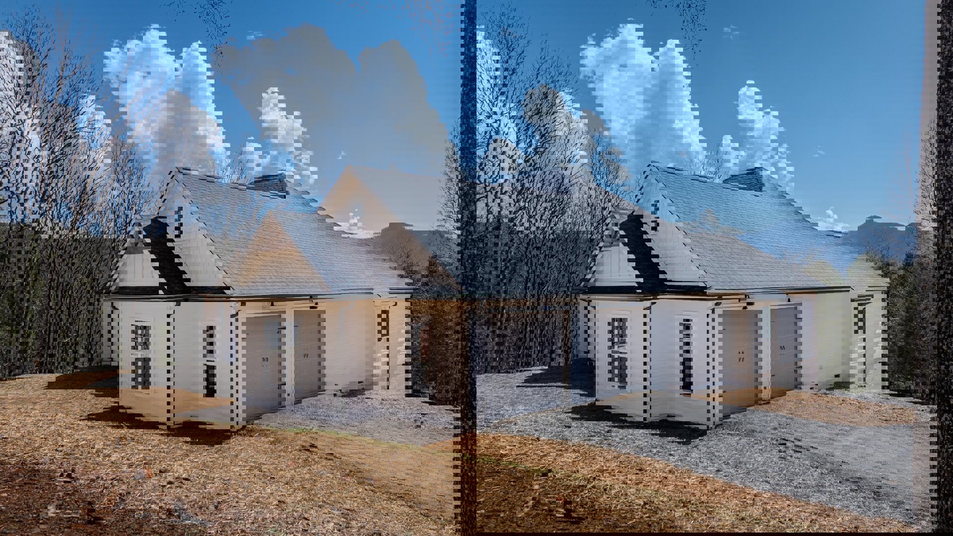 Two-story home with three white garage doors, gravel driveway, white-framed windows, gray shingle roof, and mature trees under a partly cloudy blue sky