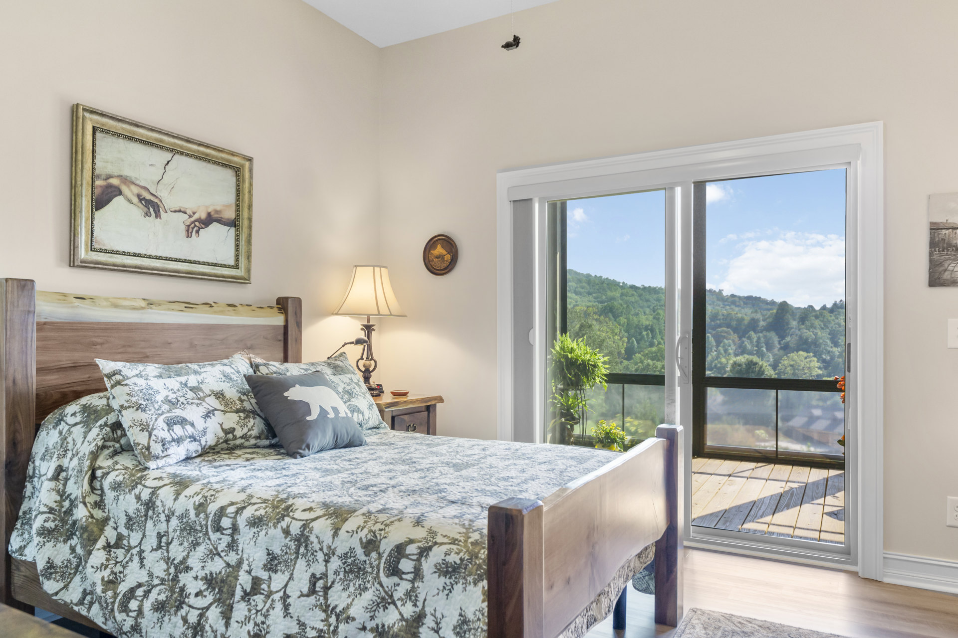 Bedroom with upholstered bed, bear motif pillow, bedside table with lamp, framed artwork of hands, glass door, and mountain landscape picture on wall