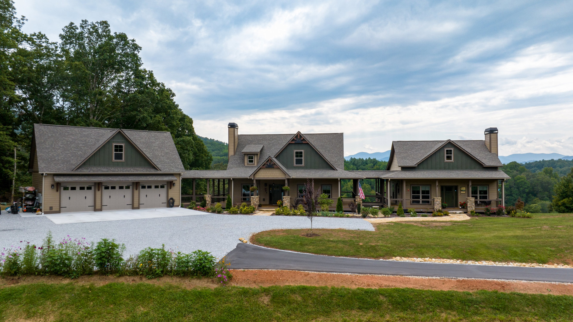 Two-story house with stone chimney, gray shingle roof, attached garage, concrete driveway, manicured lawn, and mature trees in the background