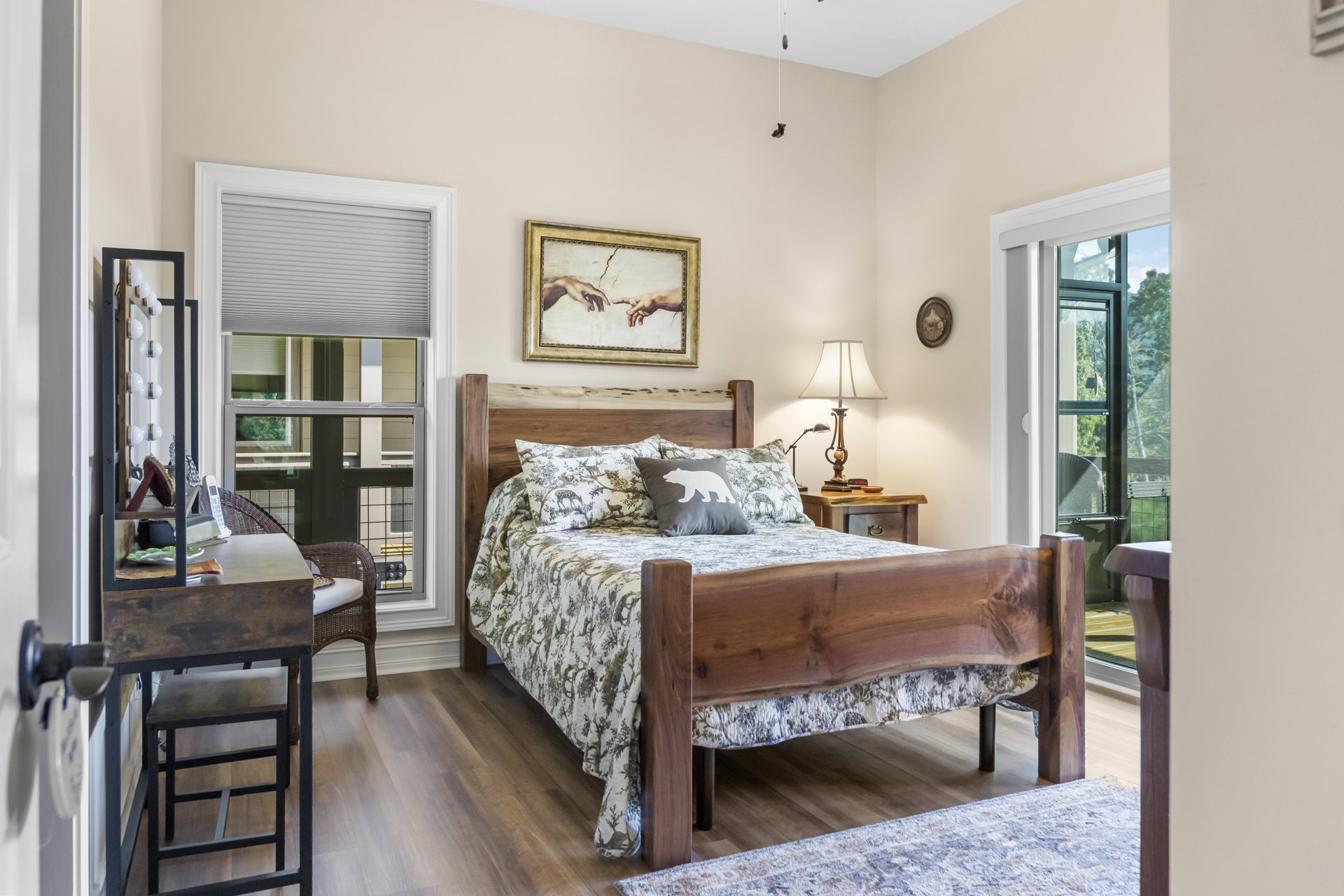 Bedroom with a wood-framed bed, white-shaded lamp on a nightstand, framed painting of hands on the wall, and a pillow featuring a bear motif