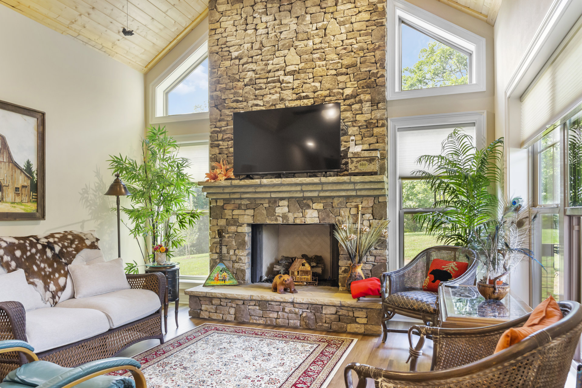 Living room with stone fireplace and mounted television, light-colored couch on patterned rug, armchair in foreground, painting of barn and tree above mantel, large window with