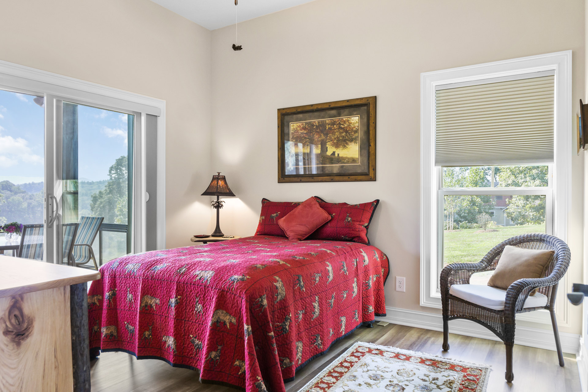 Queen bed with red blanket and matching pillows, palm tree lamp on wooden nightstand, wicker chair with patterned pillow, framed cat artwork on wall, neutral walls and carpet