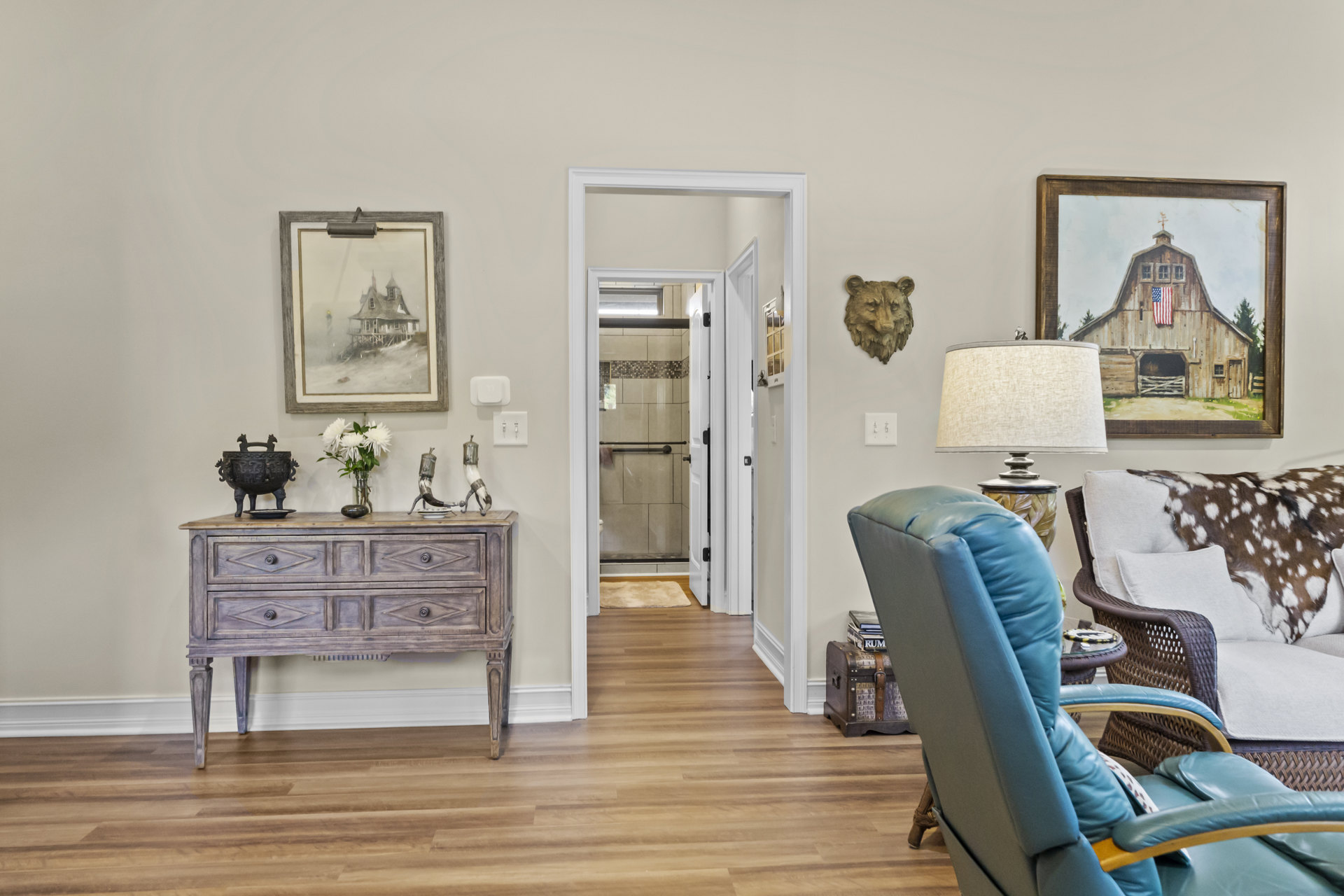 Living room with hardwood floors, blue accent chair, wooden dresser topped with lamp, cowhide draped over sofa, barn painting with flag on wall, picture frame and mirror above
