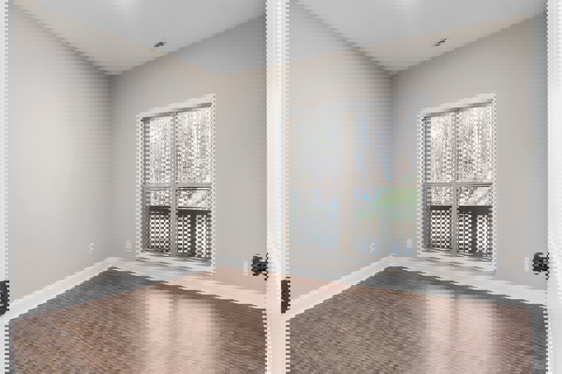 Sunlit room with large window overlooking trees and fence, hardwood floor with white baseboard trim, close-up of metal door knob and wooden railing