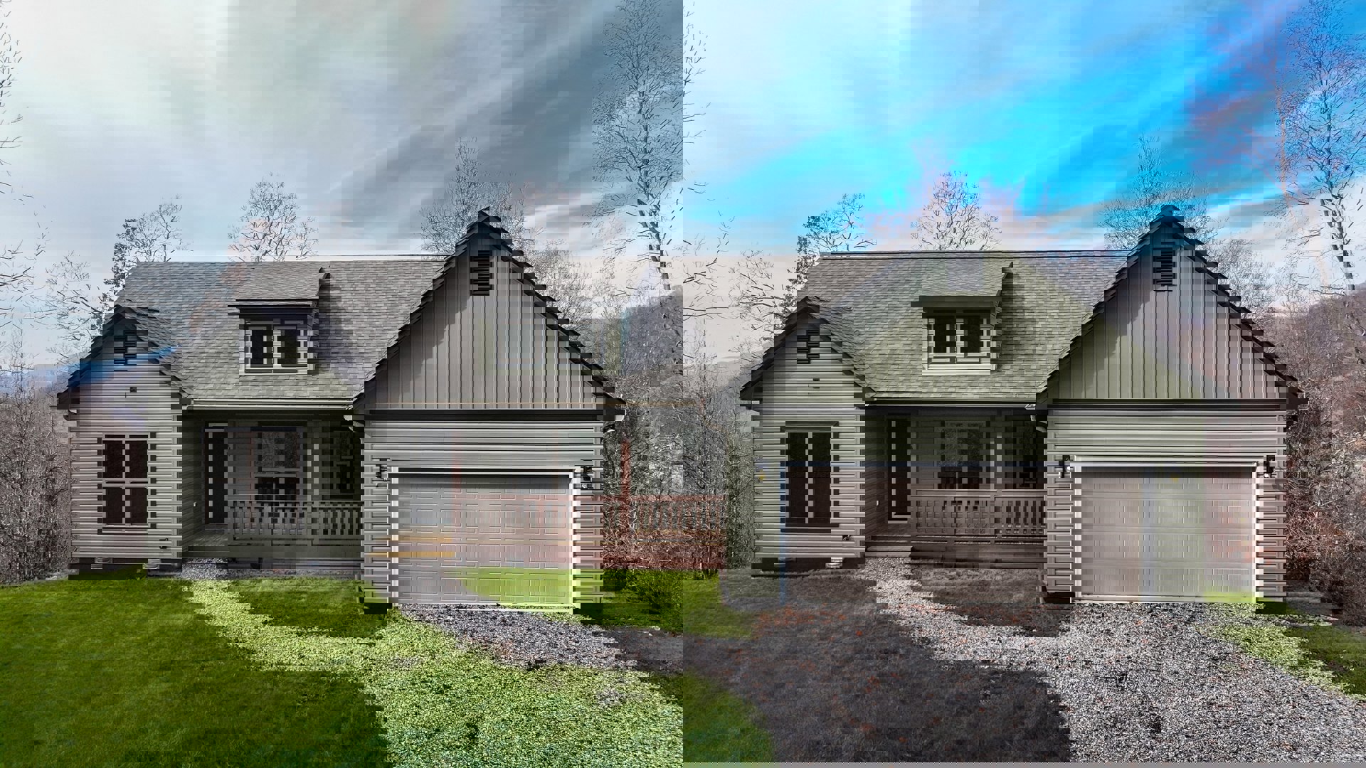 Two-story home with gray siding, white trim windows, attached garage, covered porch with wooden railing, concrete driveway, and landscaped front yard with grass and plants.