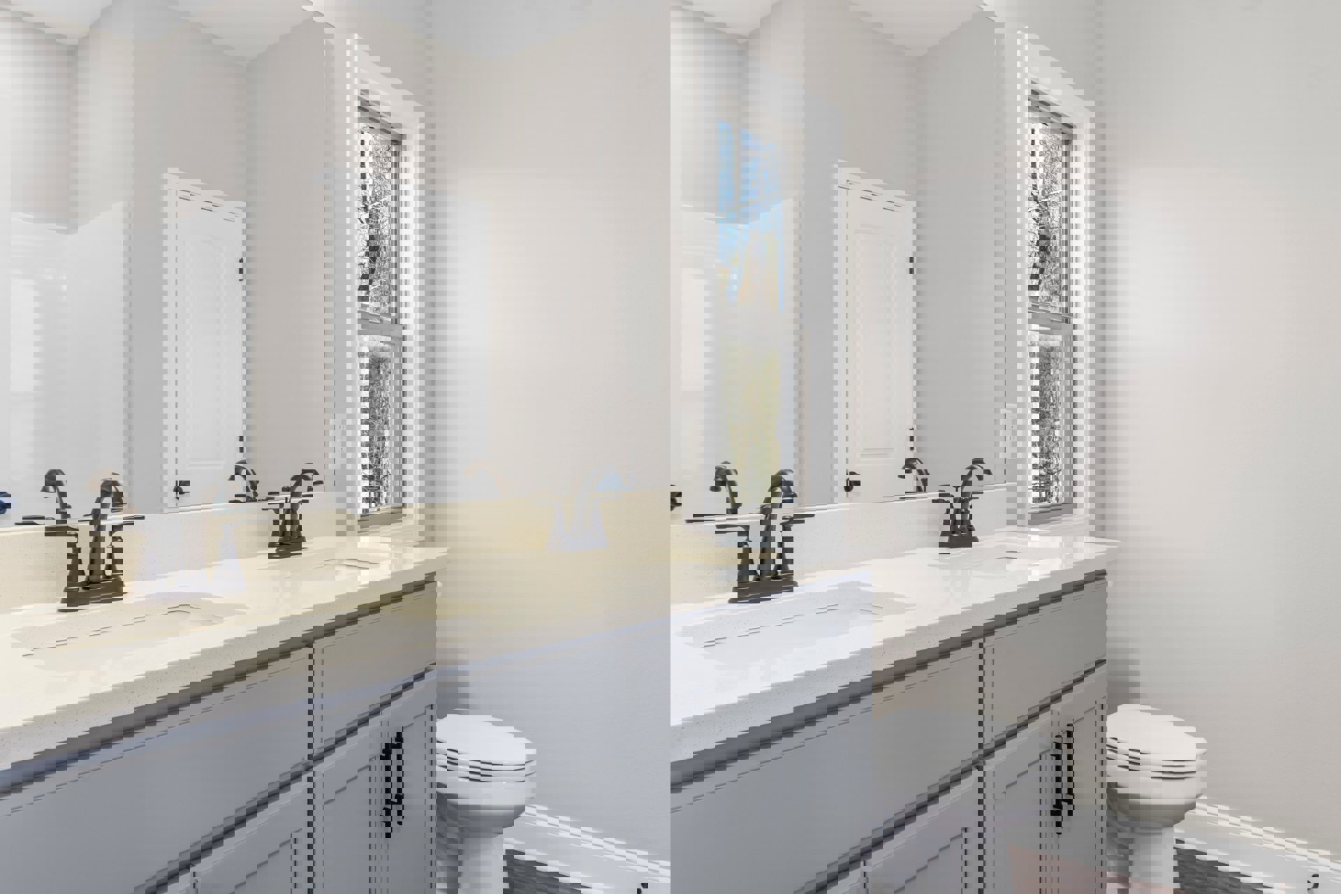 Bathroom with white toilet, white vanity sink with chrome faucet, black hardware on white door, large window showing green trees, light tile flooring, neutral walls.