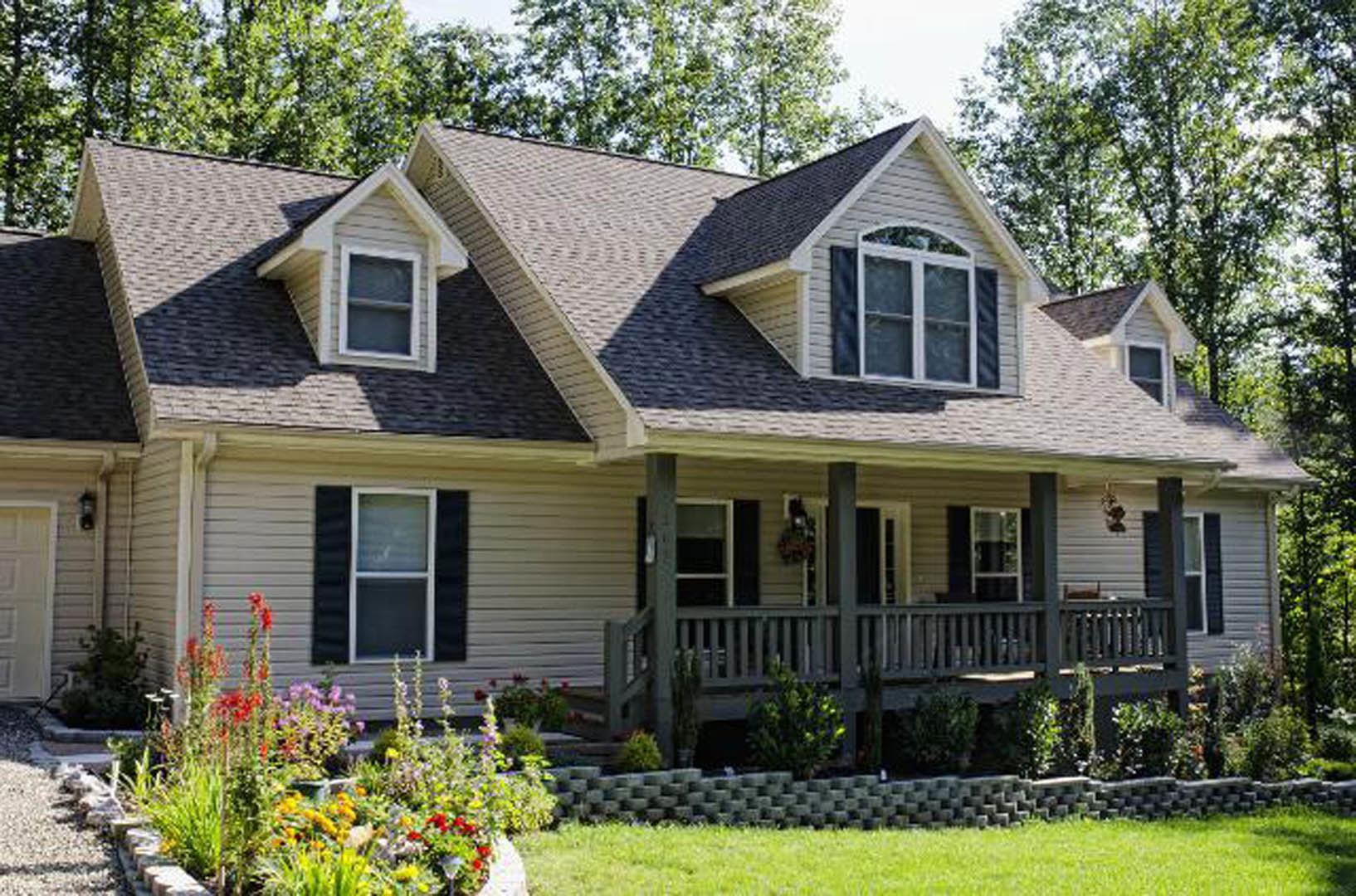 White siding house with black shuttered windows, covered front porch with white railings, flower garden and green lawn in the front yard.