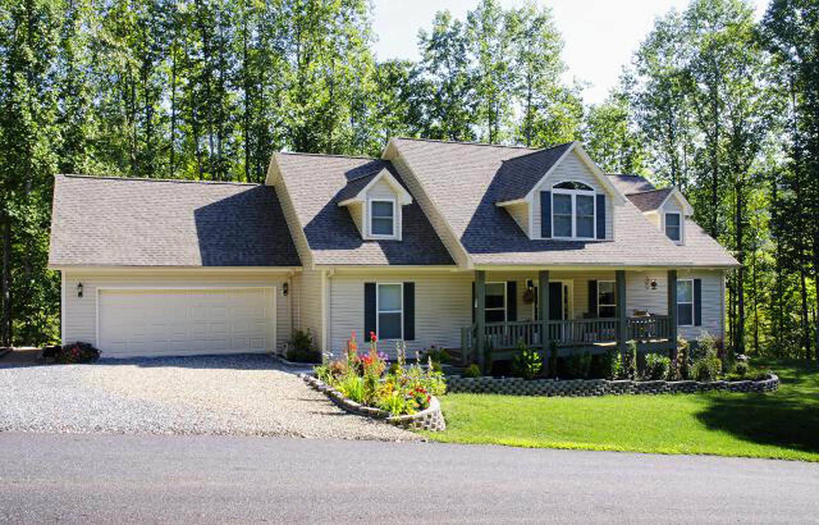 Two-story house with white siding and blue trim, attached garage with white door, flower bed along front porch, paved driveway, mature trees in background