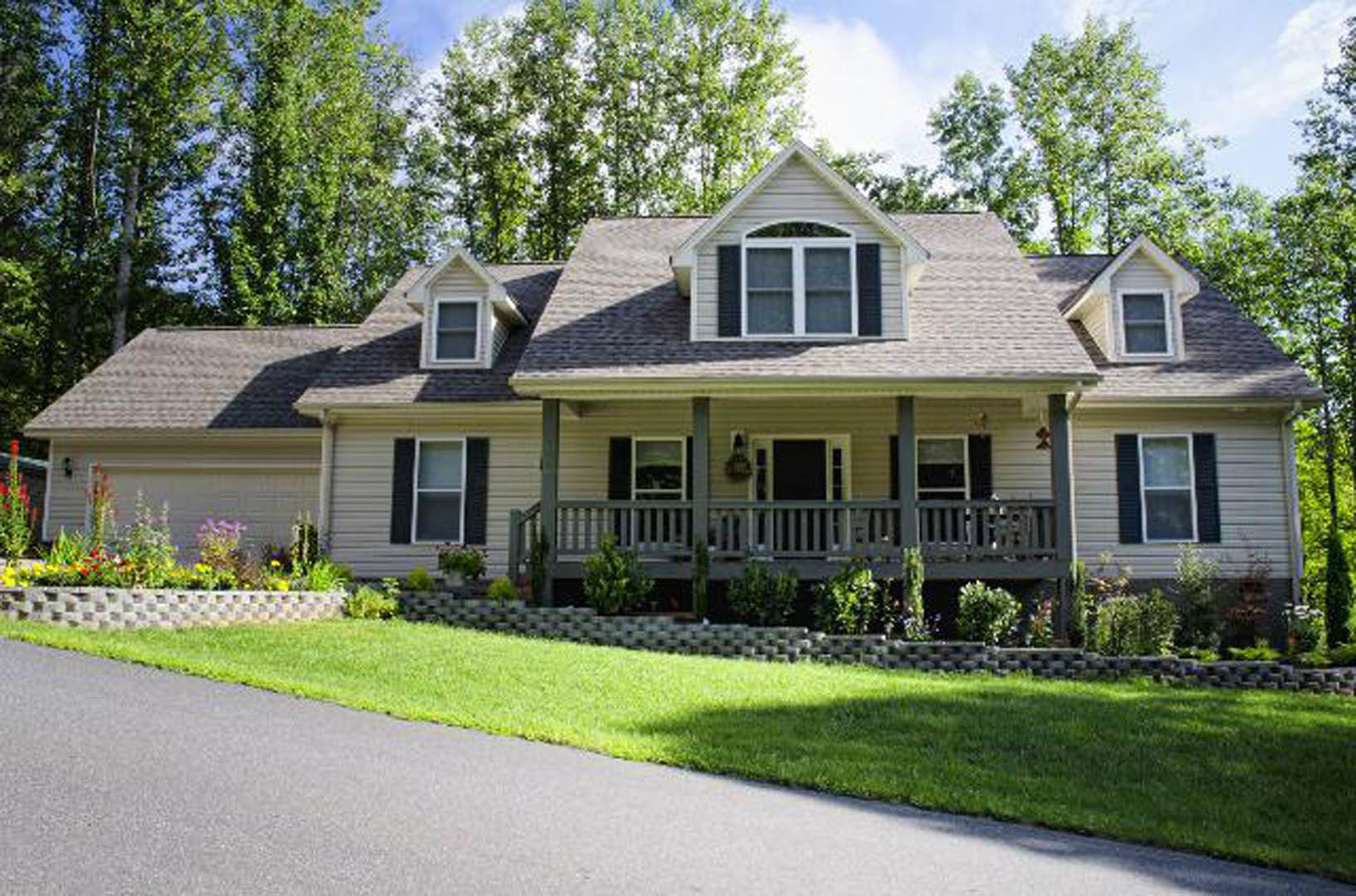 Two-story house with white siding, covered front porch, paved driveway, manicured lawn, large windows with white frames, and mature trees in the yard