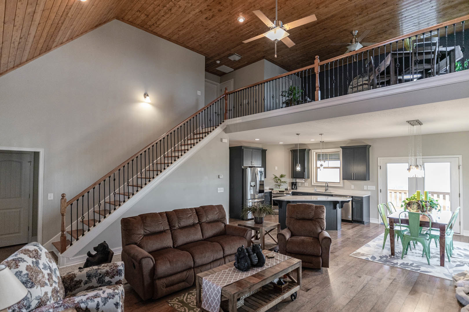 Open concept living room with brown couch and floral patterned sofa, coffee table featuring black bear figurines, adjacent kitchen area, and prominent large staircase with wood