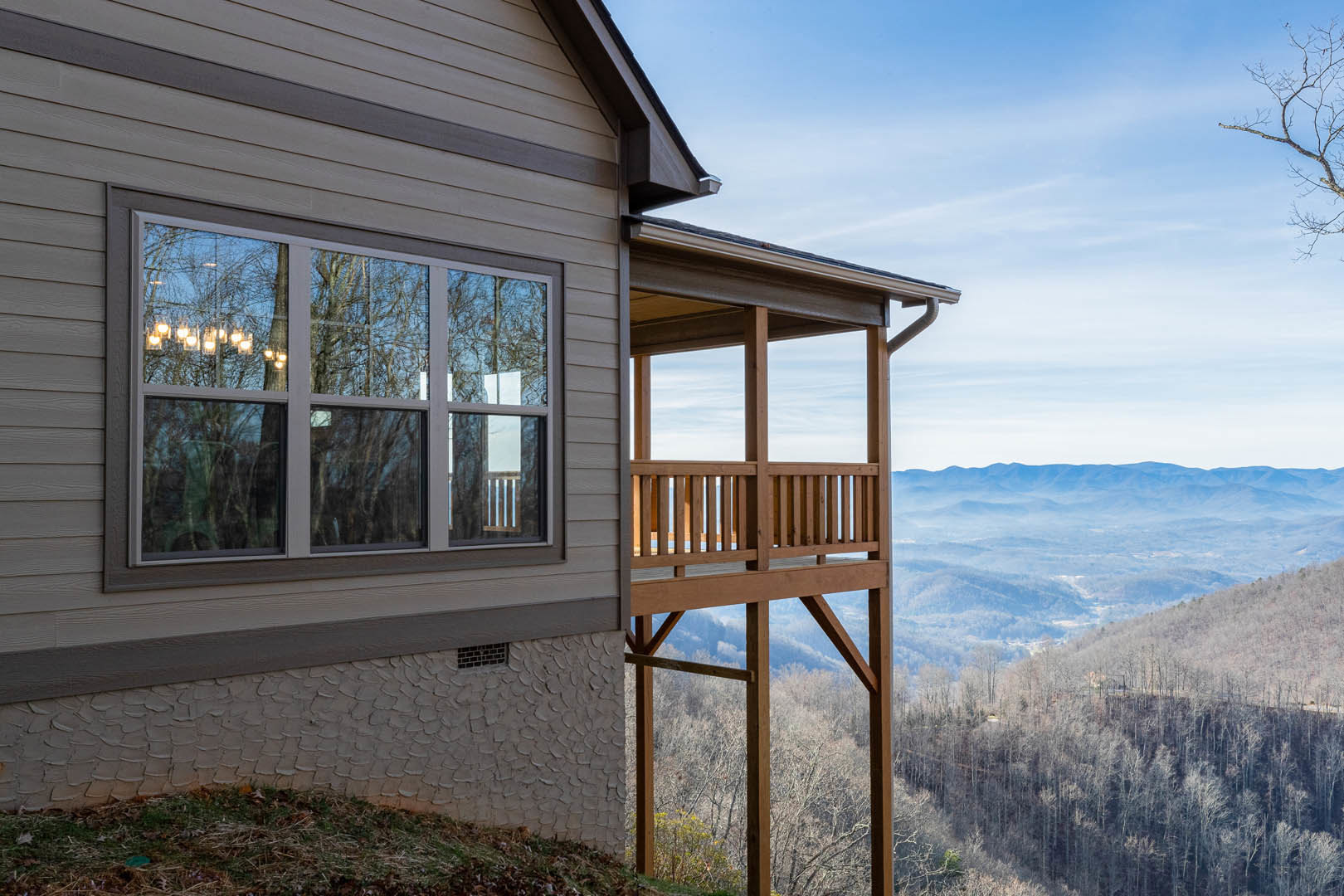 Modern home with wooden balcony, large windows, and light fixture, overlooking lush green valley and dense forest under cloudy sky