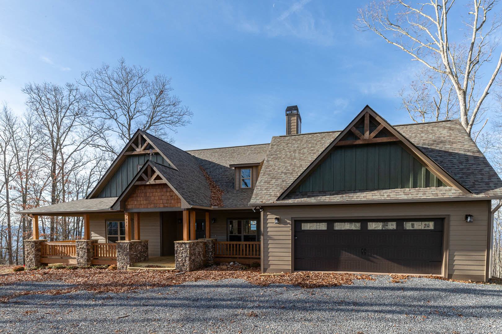 Two-story house with gray siding, attached garage with white paneled door, gravel driveway scattered with leaves, wooden porch railing, multi-pane windows, and small balcony on