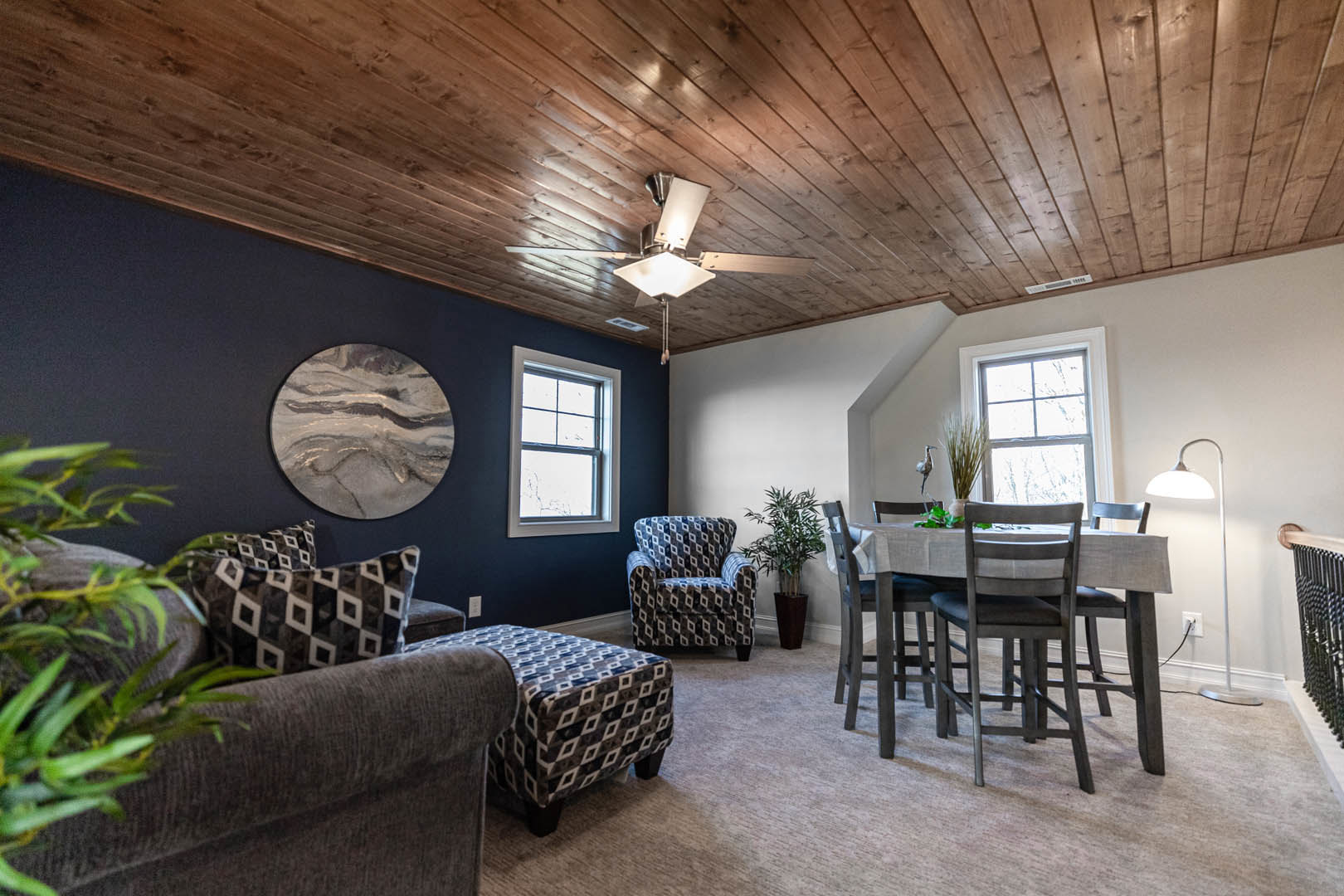 Living room with wood table and upholstered chairs, blue and white patterned accent chair, white-framed window, potted plant, neutral walls, hardwood floor, circular wall decor