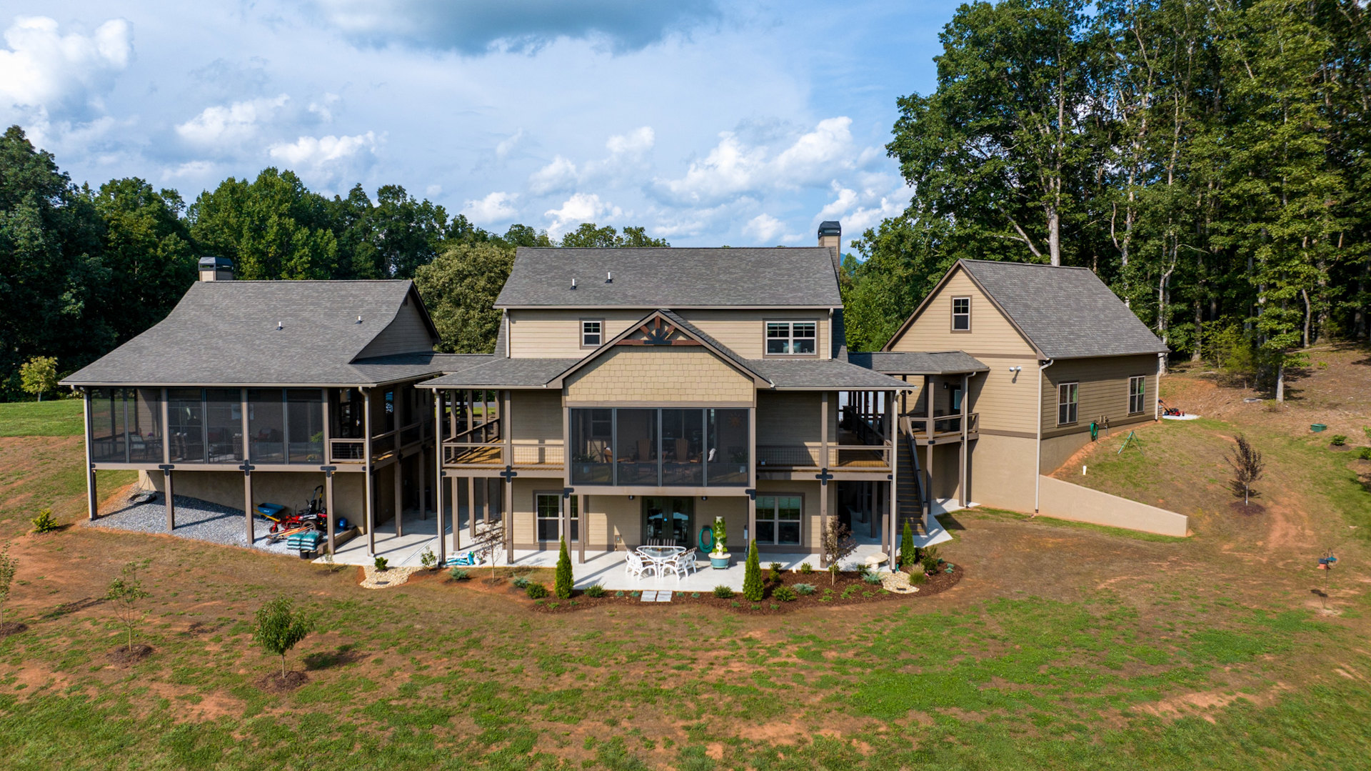 Two-story house with light siding, covered porch, manicured lawn, mature trees, and blue sky with scattered clouds