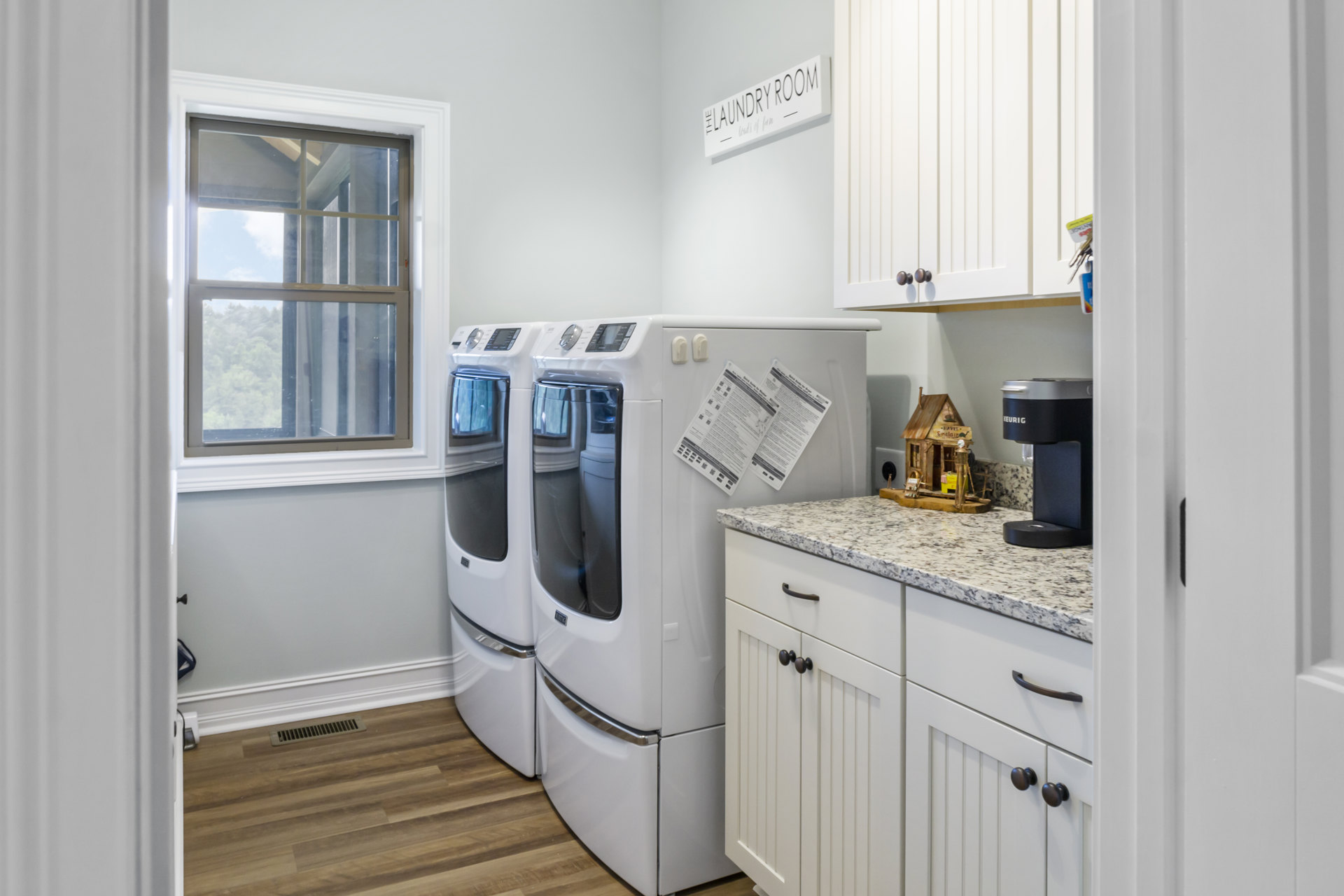 Laundry room featuring white washer and dryer, white cabinetry, countertop, window overlooking trees and sky, wall sign, and paper with text.