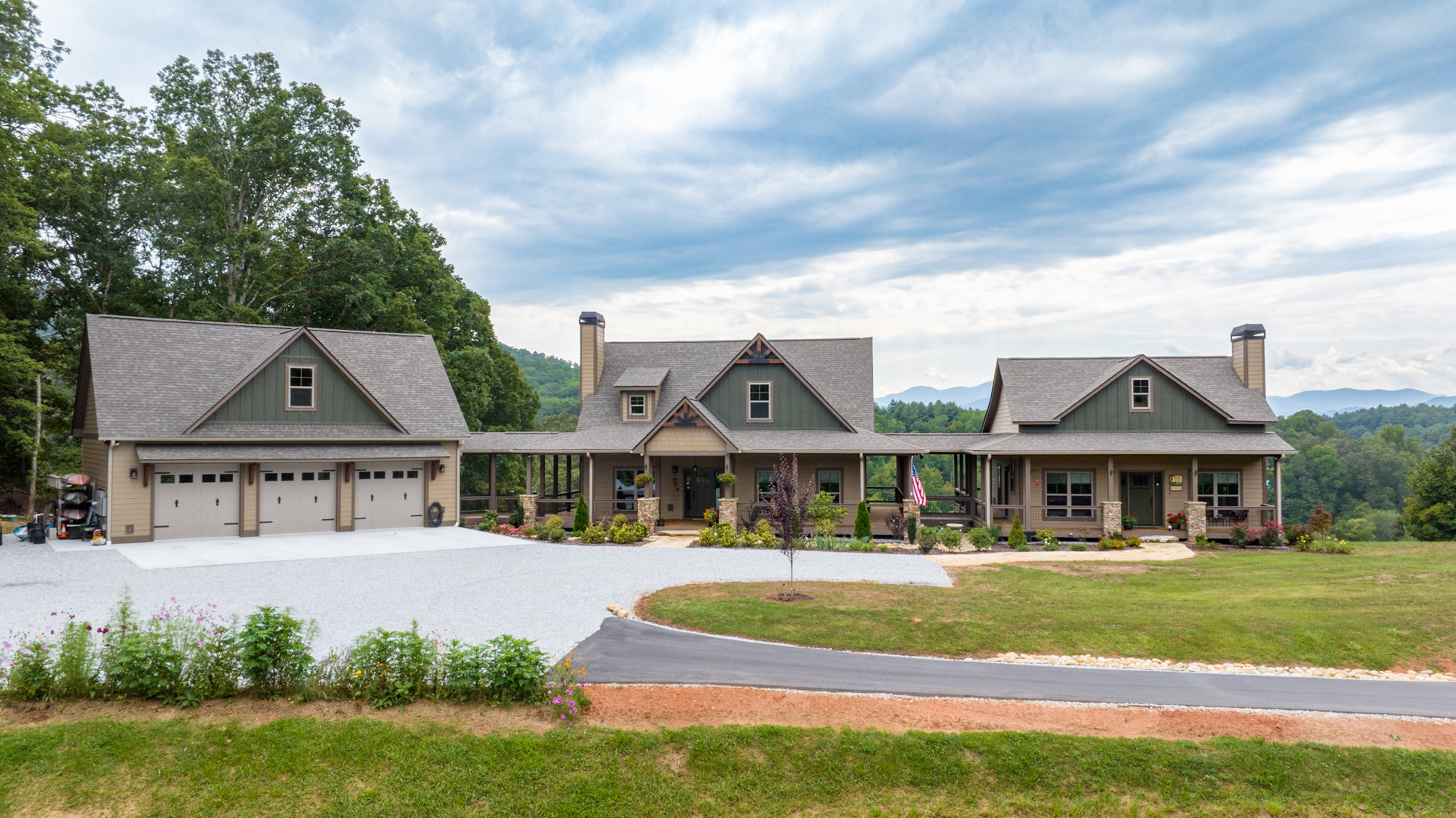 Two-story house with gray siding, shingled roof, attached garage, gravel driveway bordered by green grass, purple flowers in window box, chimney, and mature trees in background