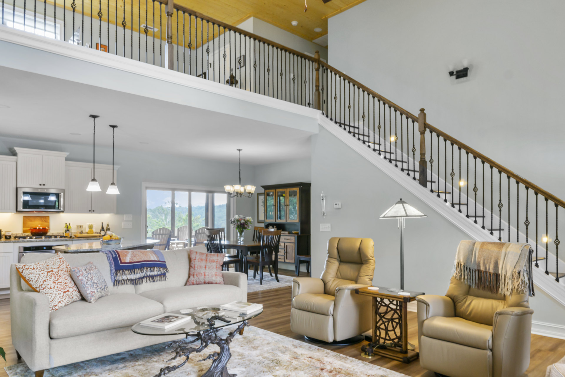 Living room with hardwood floors, neutral couch with patterned pillows, tan recliner chair draped with a blanket, staircase with wooden handrail, white walls, and soft ambient