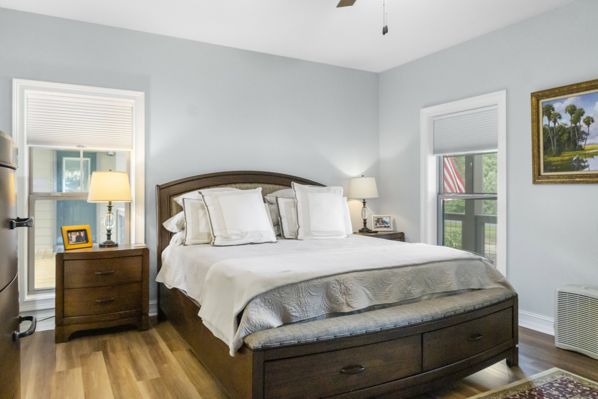 Queen bed with natural wood frame, white bedding, ceiling fan above, nightstand with white-shaded lamp, bird painting on wall, dresser topped with framed photo