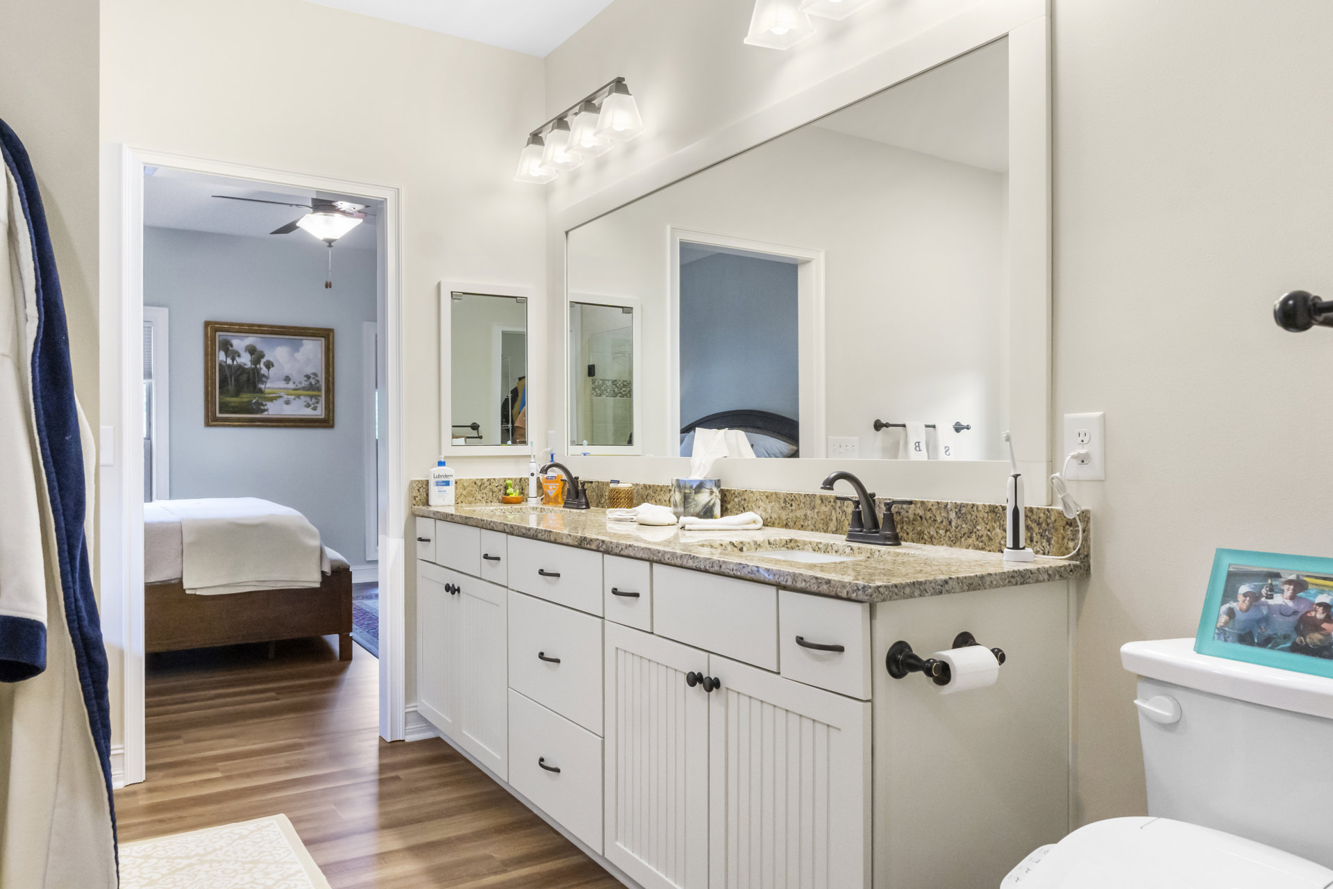Bathroom with double sink vanity, expansive wall mirror, white cabinetry, chrome faucets, decorative gold-framed landscape painting, blue and white blanket folded nearby, framed