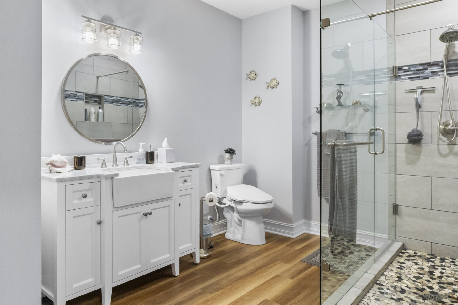 Bathroom with tiled shower, white toilet and toilet paper holder, round silver-framed mirror above white cabinet with silver knobs, pair of white metal fixtures, and a blurry fish