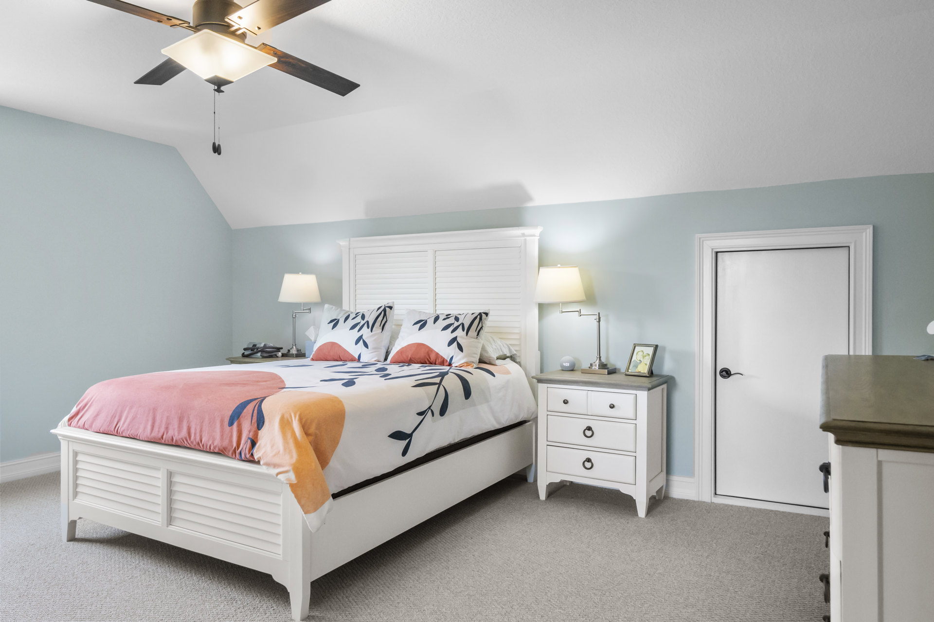 Bedroom with white door, white bed frame, white dresser topped with picture frames, lamp with white shade, and neutral walls.