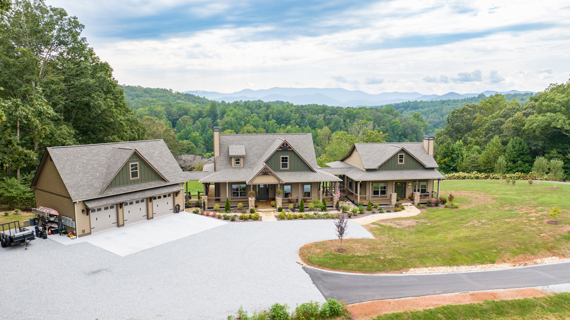 Two houses with pitched roofs, connected by a shared driveway, surrounded by green lawns and mature trees under a partly cloudy blue sky; one house features an attached garage, and