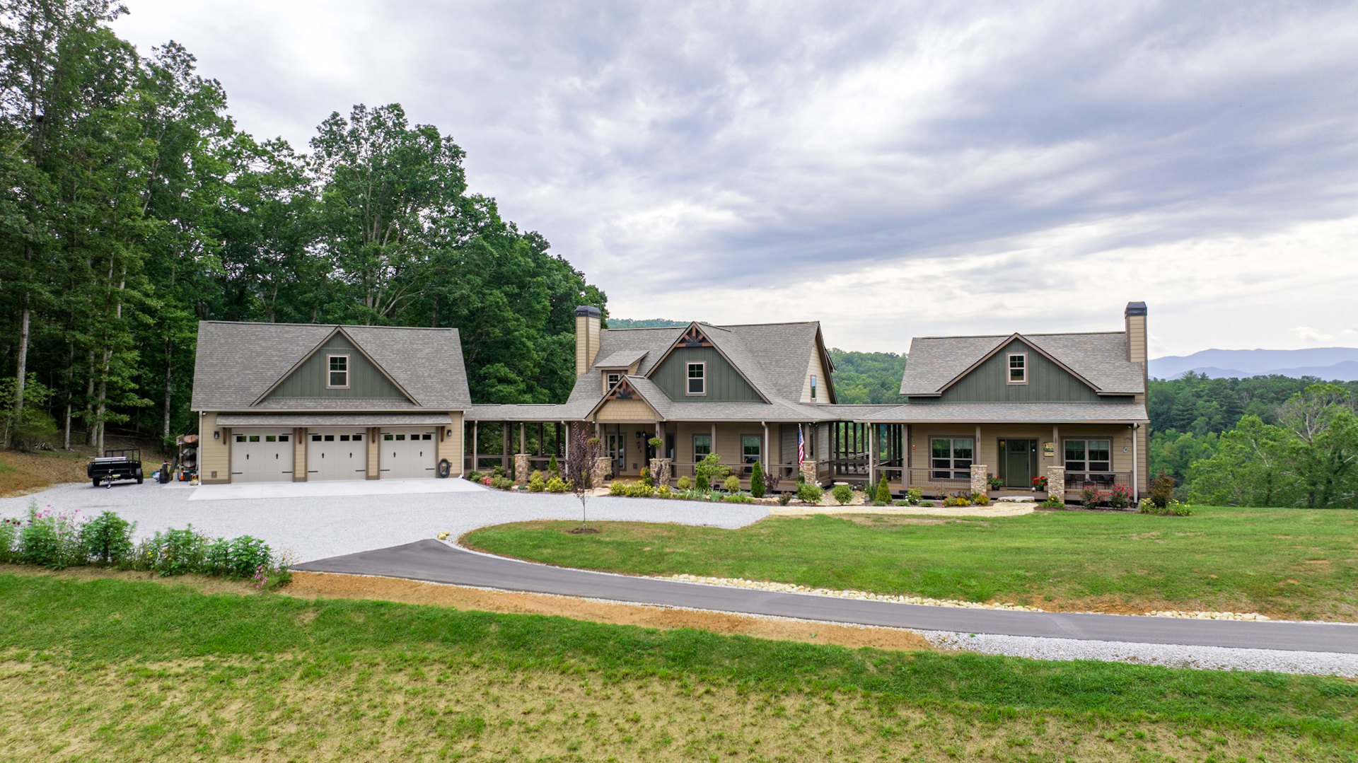 Two-story house with white-framed windows, expansive front porch, paved driveway, mature trees, manicured grass, and a row of bushes bordering a pond.