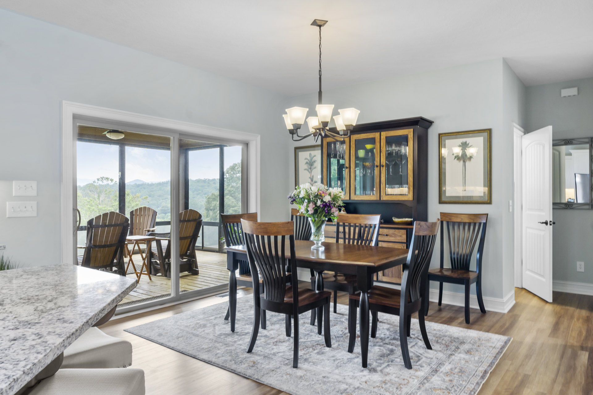 Dining room with wooden table and upholstered chairs, glass vase with green leaves centerpiece, large glass doors, framed palm tree artwork on wall, light hardwood flooring, white