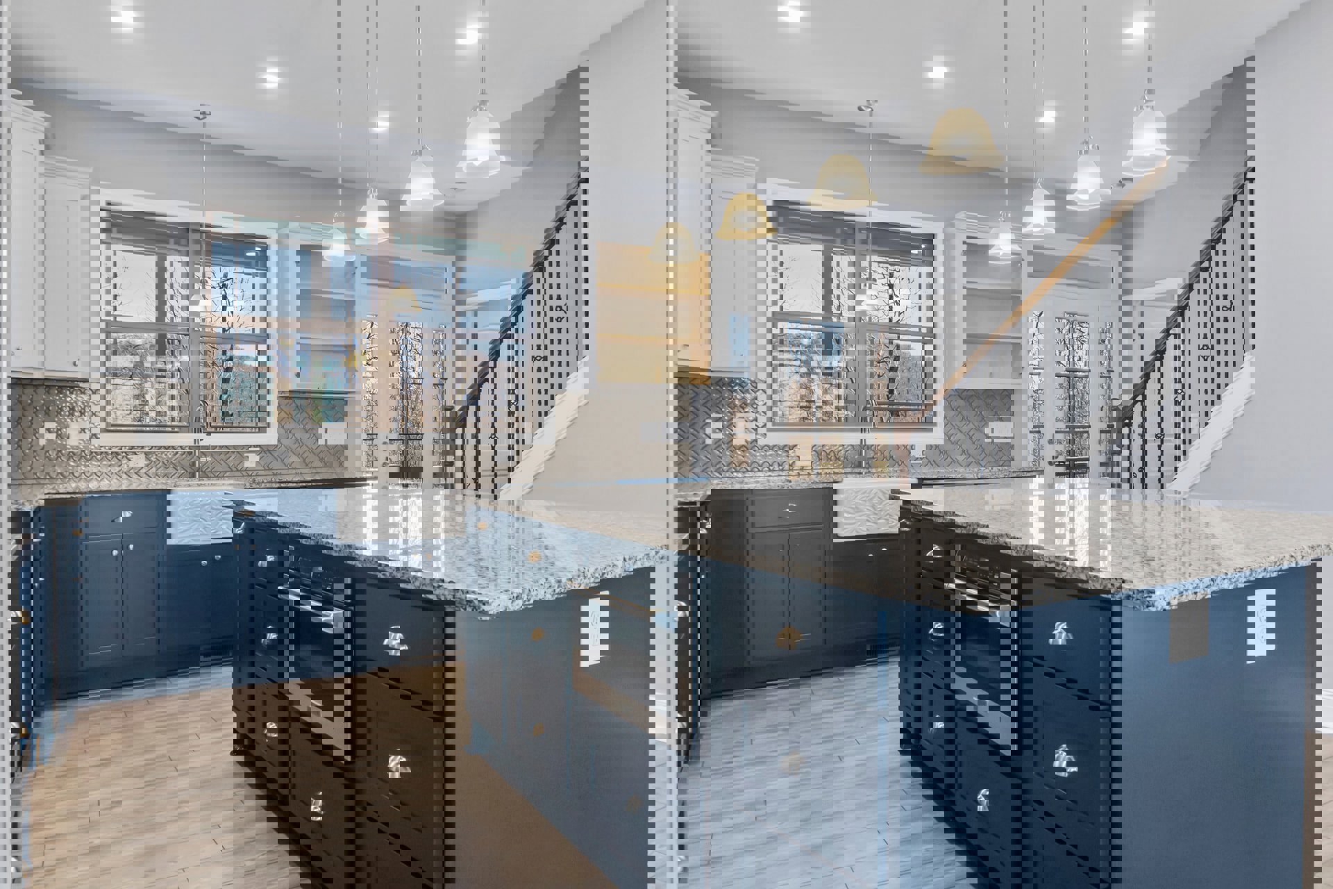 Marble kitchen island with white cabinetry, stainless steel oven, sink beneath large windows, wood flooring, blue door, staircase with modern light fixture, and white electrical