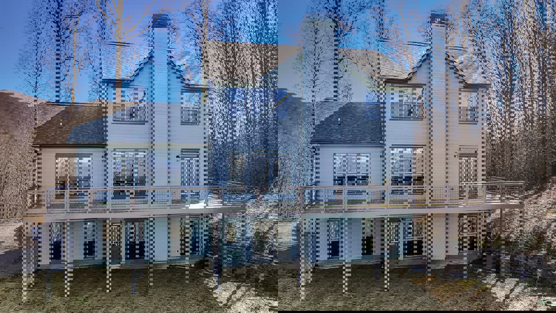 Two-story home with white-framed windows, covered front porch, upper balcony, wooden deck, and expansive grassy yard bordered by mature trees