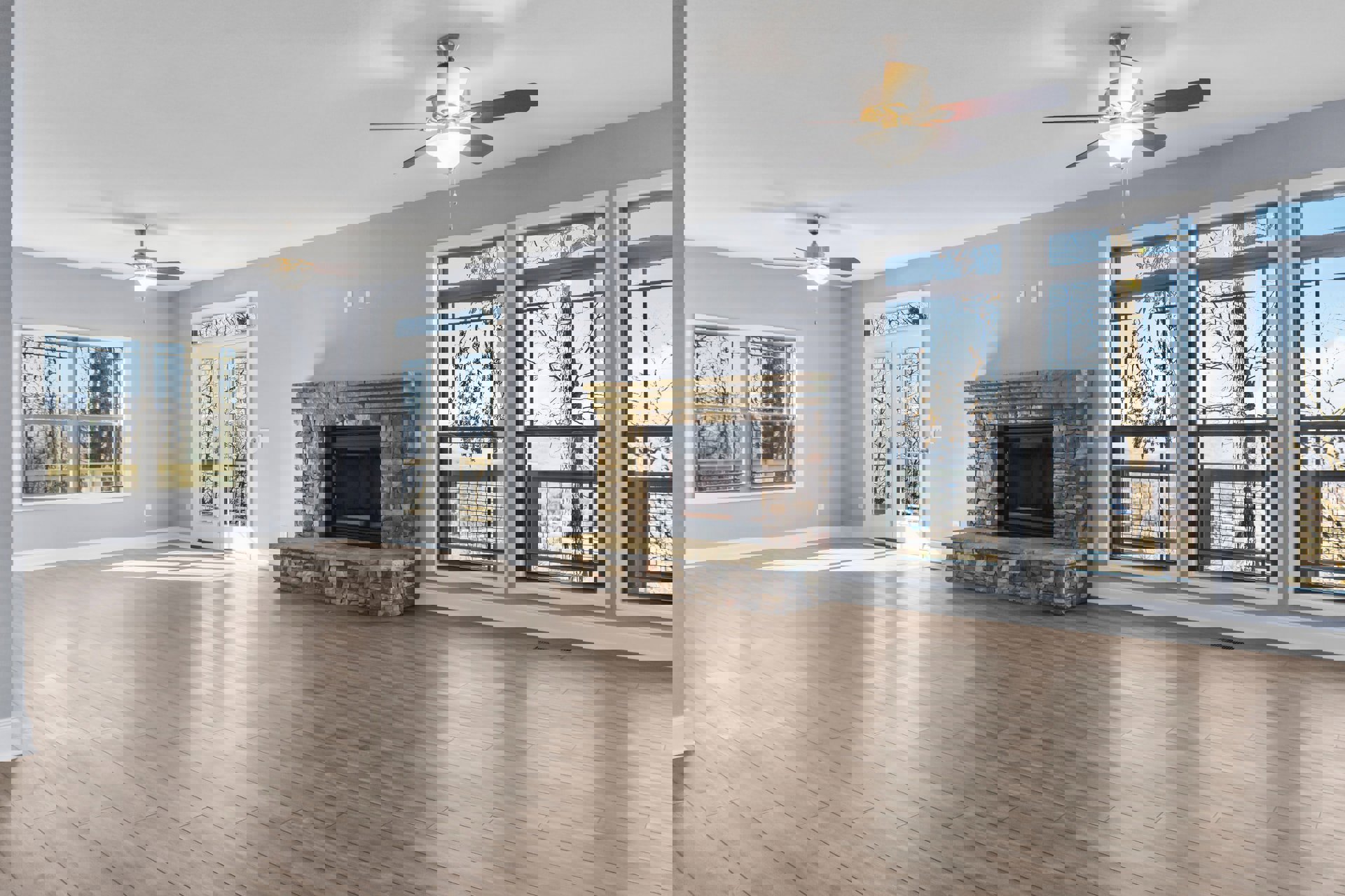 Living room with wood flooring, ceiling fan, stone fireplace, and large windows framing views of tree trunks and branches outside.