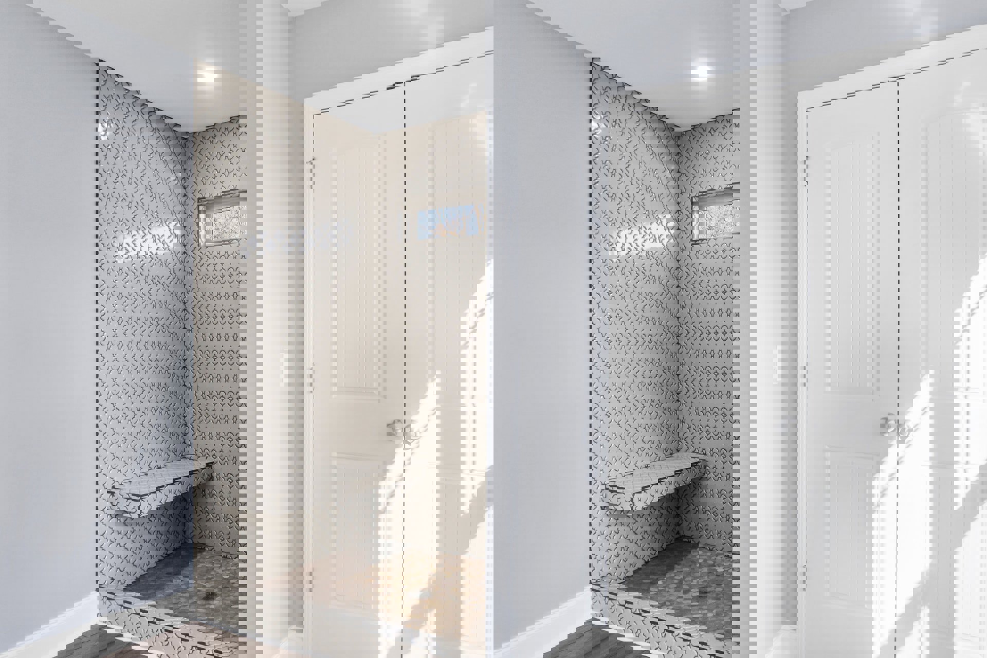 Walk-in shower with glass door, built-in bench, white tile walls featuring black patterned shelf, silver hardware, window with white frame, marble floor, and plaster ceiling.