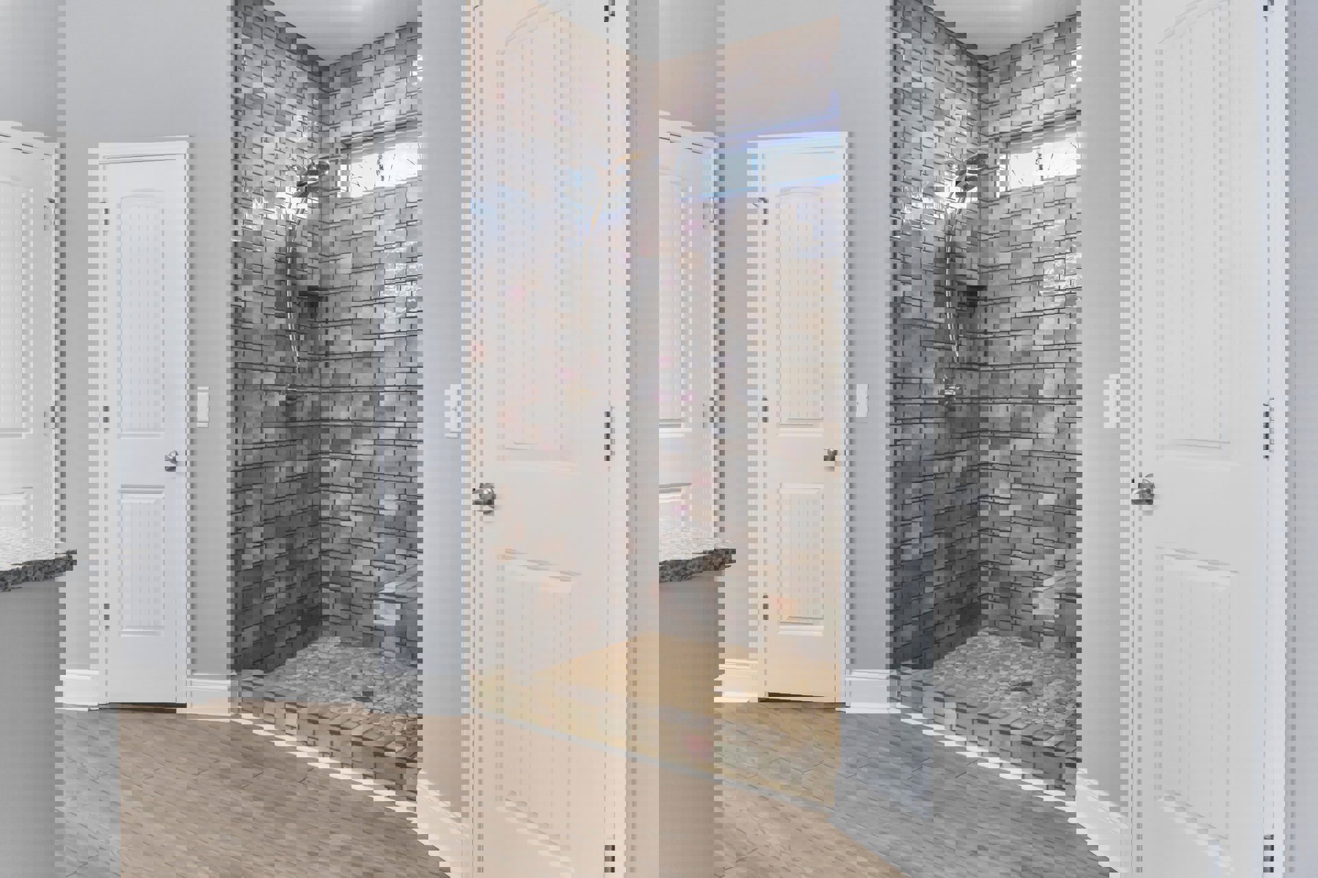 Walk-in shower with gray tile floor, glass enclosure, and chrome shower head; adjacent white door with silver doorknob; wood flooring and exposed brick wall visible in background